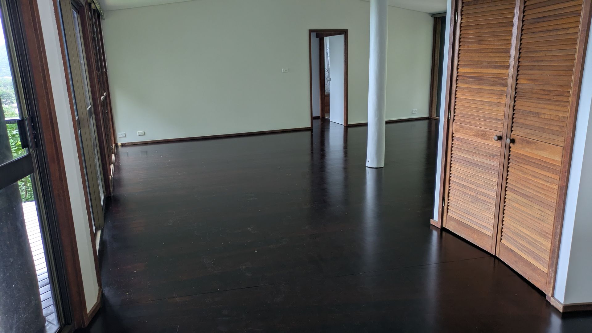 Empty room with dark wood floor, white walls, and a column. Tall, shuttered doors are on the right.