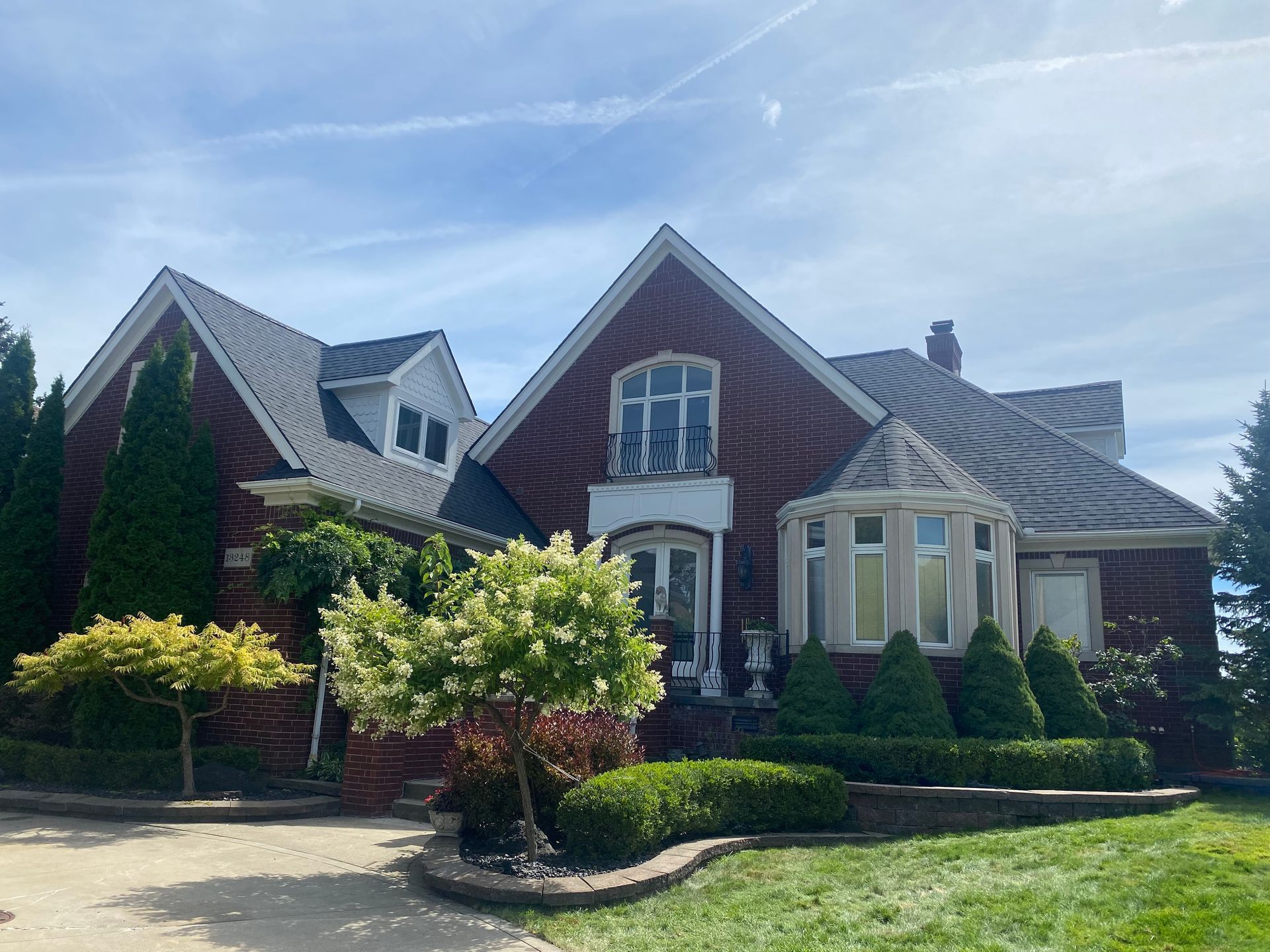 A large brick house with a gray roof is surrounded by trees and bushes.