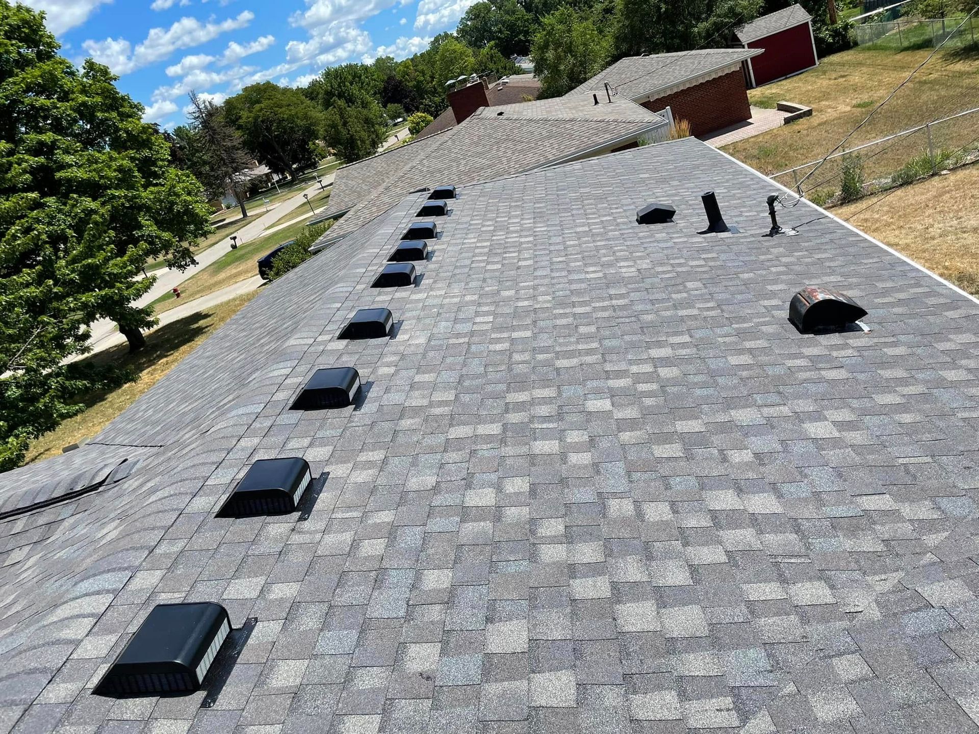 A view from above showing a grey shingled roof with a line of black ventilation covers, next to trees and a red building.