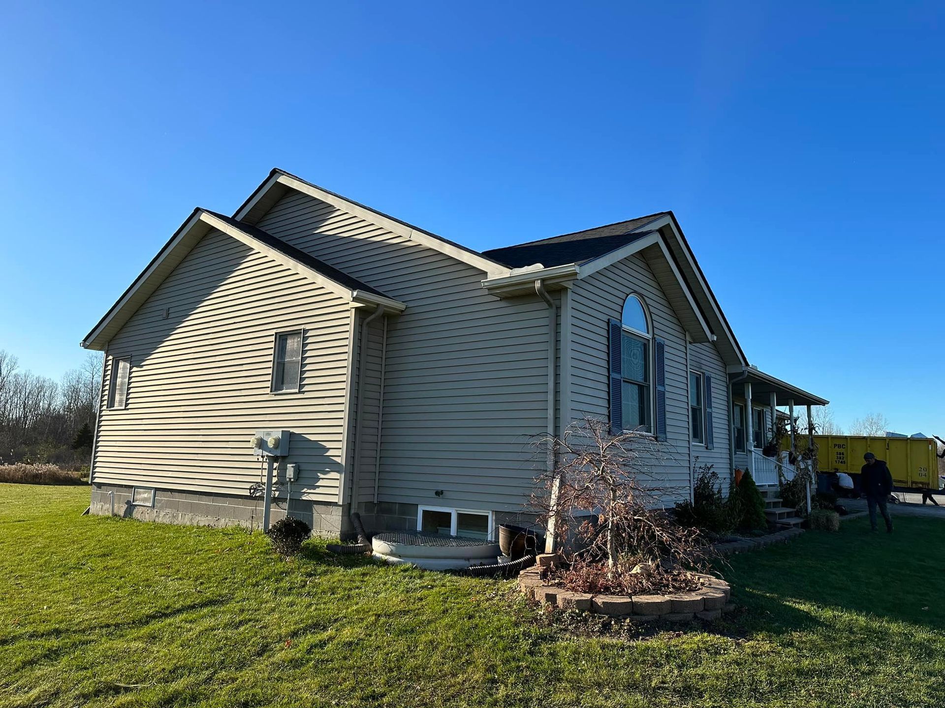 Tan house with a dark roof sits on a grassy lawn under a clear blue sky. A small bush is in front.