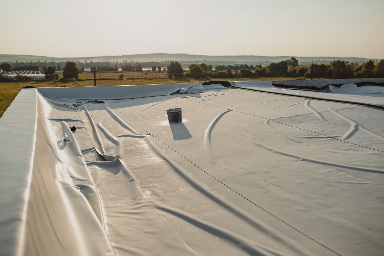 White rooftop membrane with seams and vents, viewed across a flat industrial roof toward a distant landscape.
