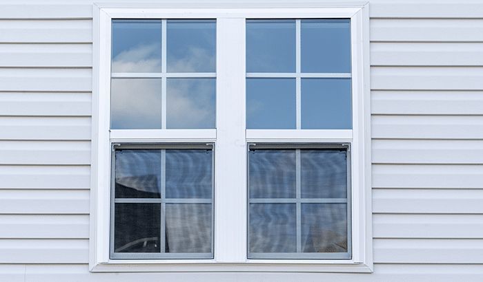 White double-hung window with a grid pattern, set in light gray siding. Reflects sky and unseen objects.