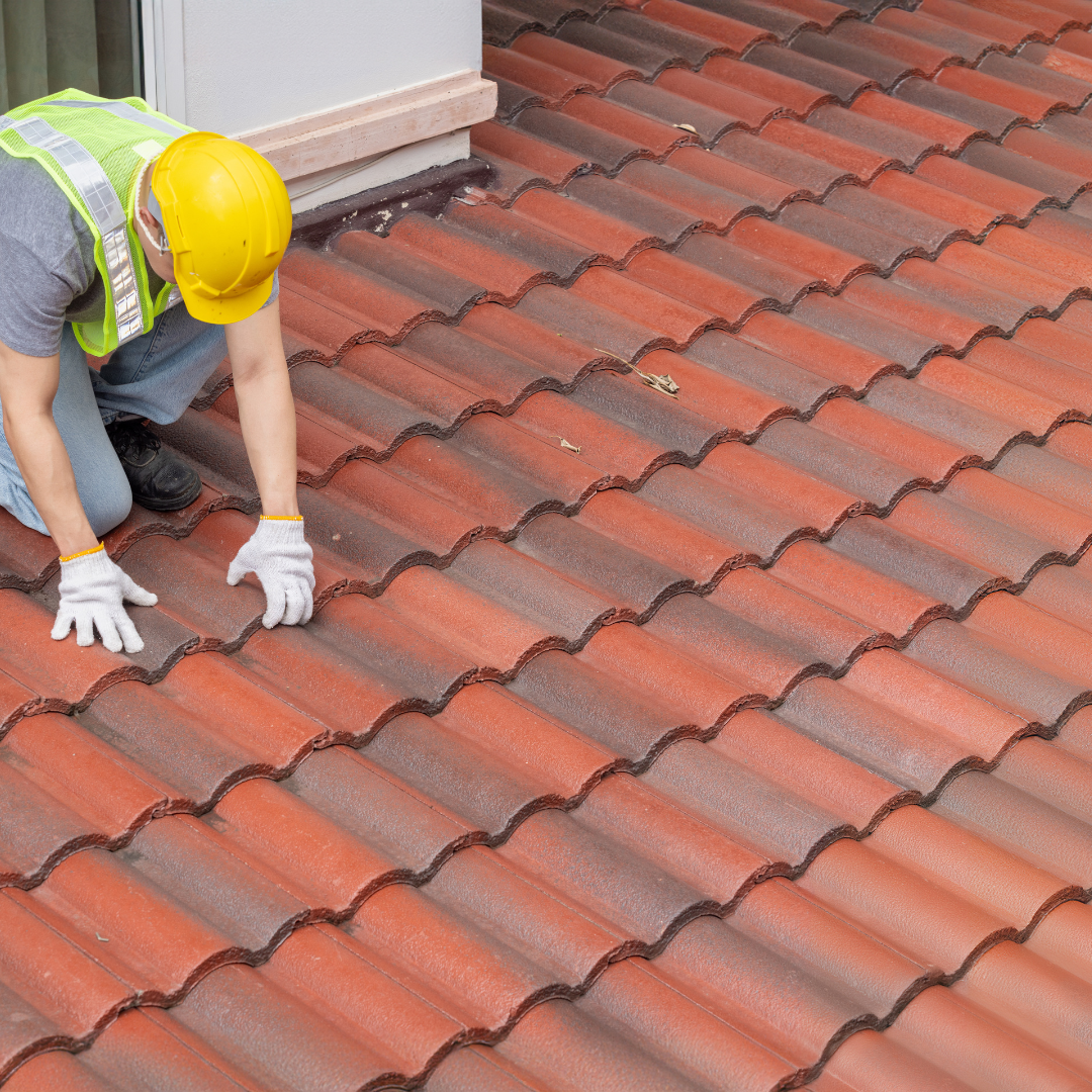 A man wearing a hard hat and safety vest is kneeling on a tiled roof.