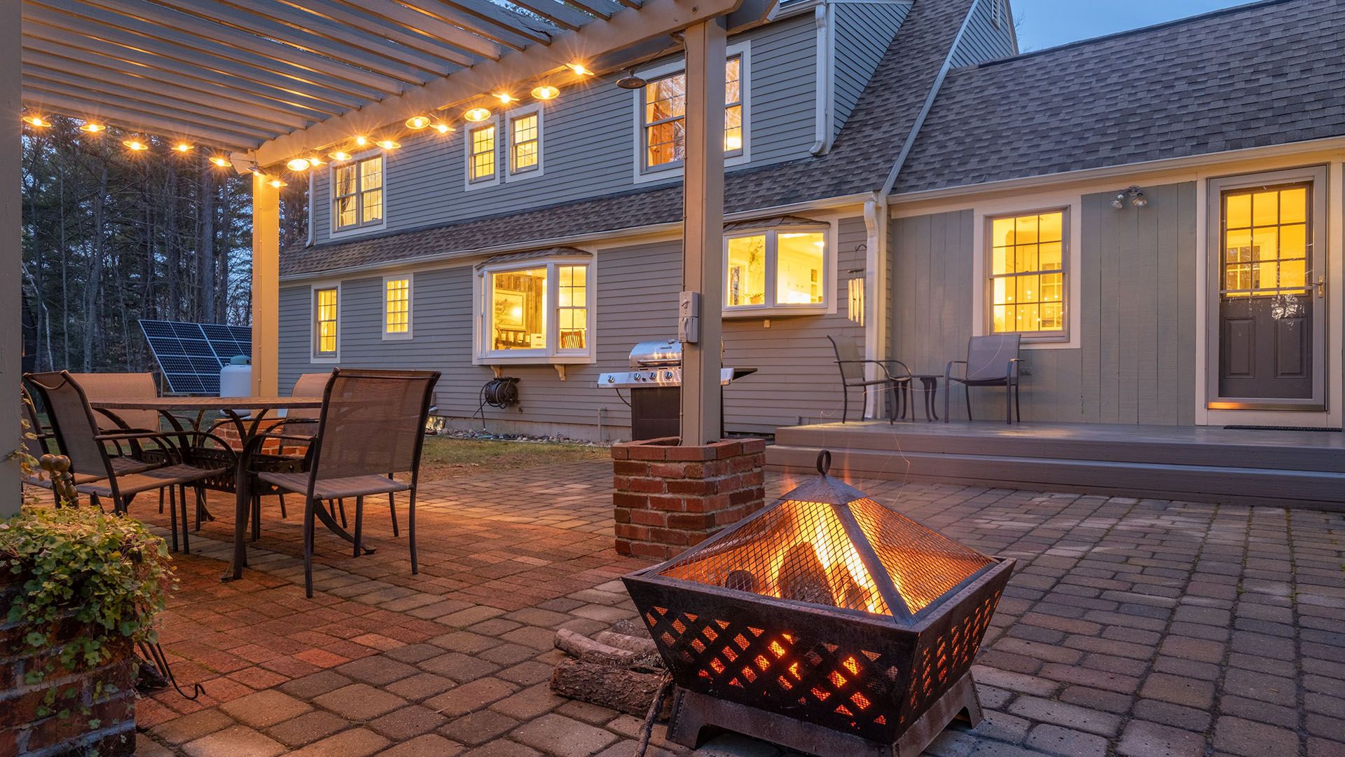 Backyard patio with fire pit, dining table, and string lights, in front of a house.