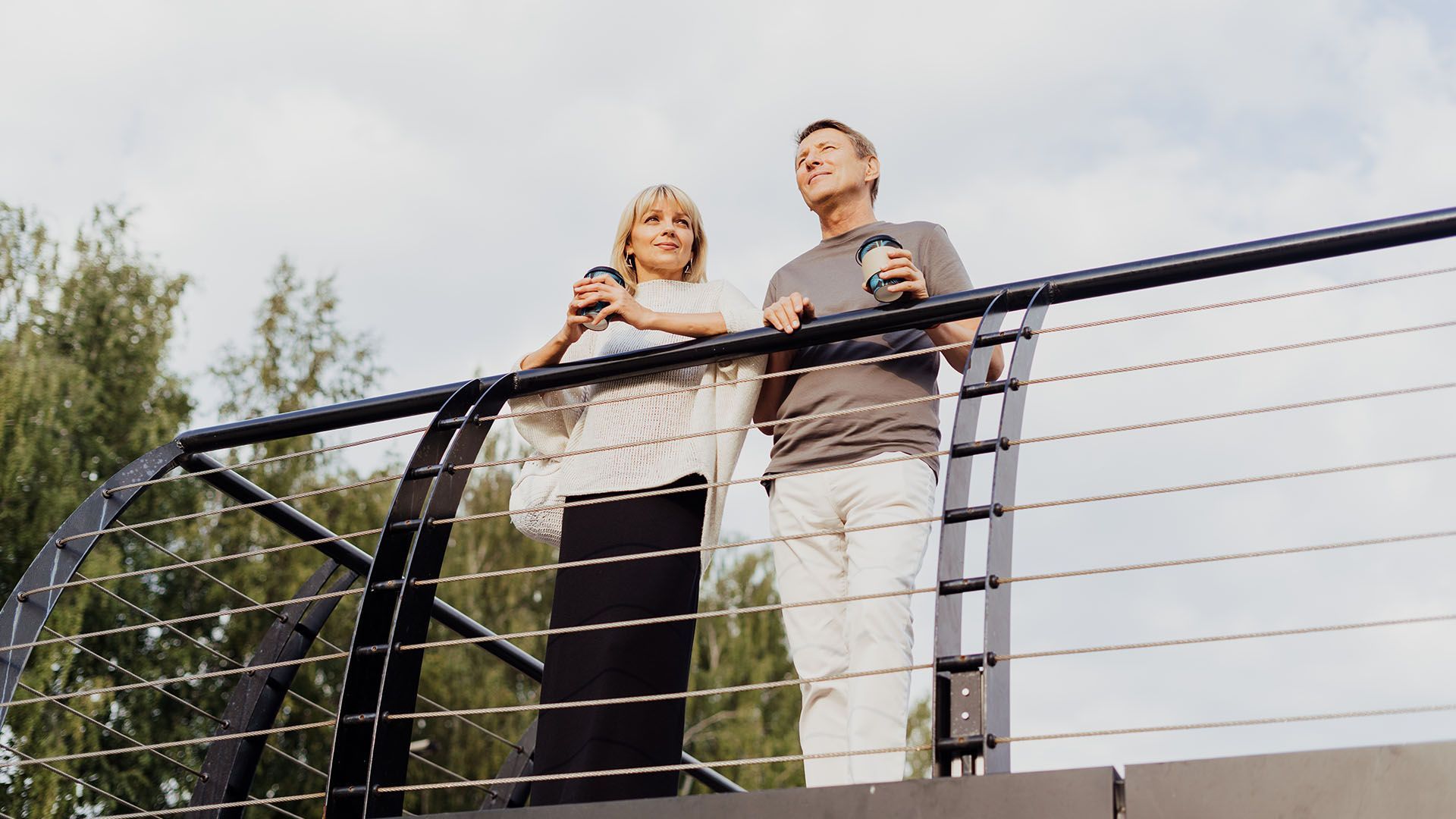 Couple on a bridge, holding mugs, looking up, cloudy sky.