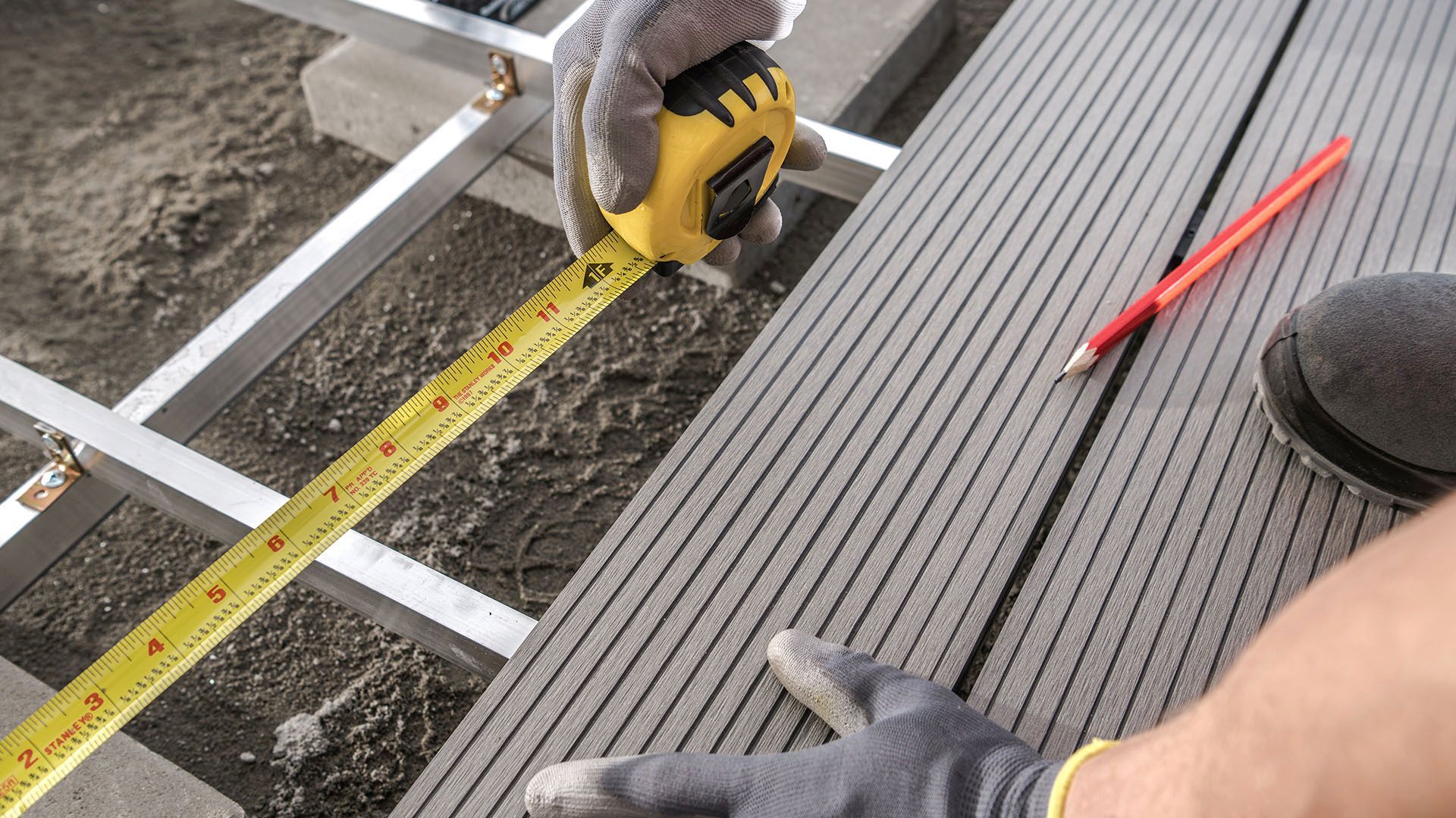 Person wearing gloves measures a gray deck board with a yellow tape measure during construction.