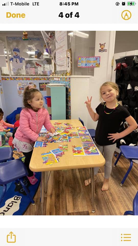 Two young girls at a table with puzzles, one gives a peace sign. Indoors, classroom setting.