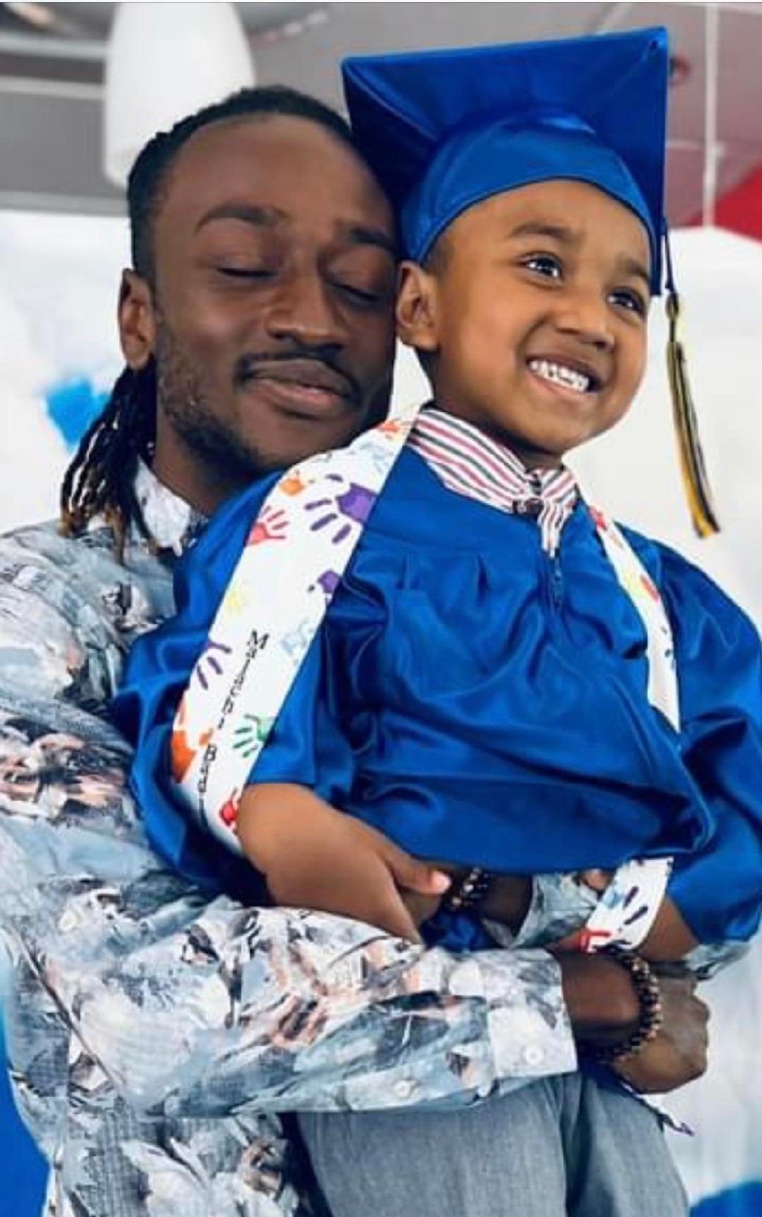 Man holding a smiling child in graduation attire; both are inside with balloons.