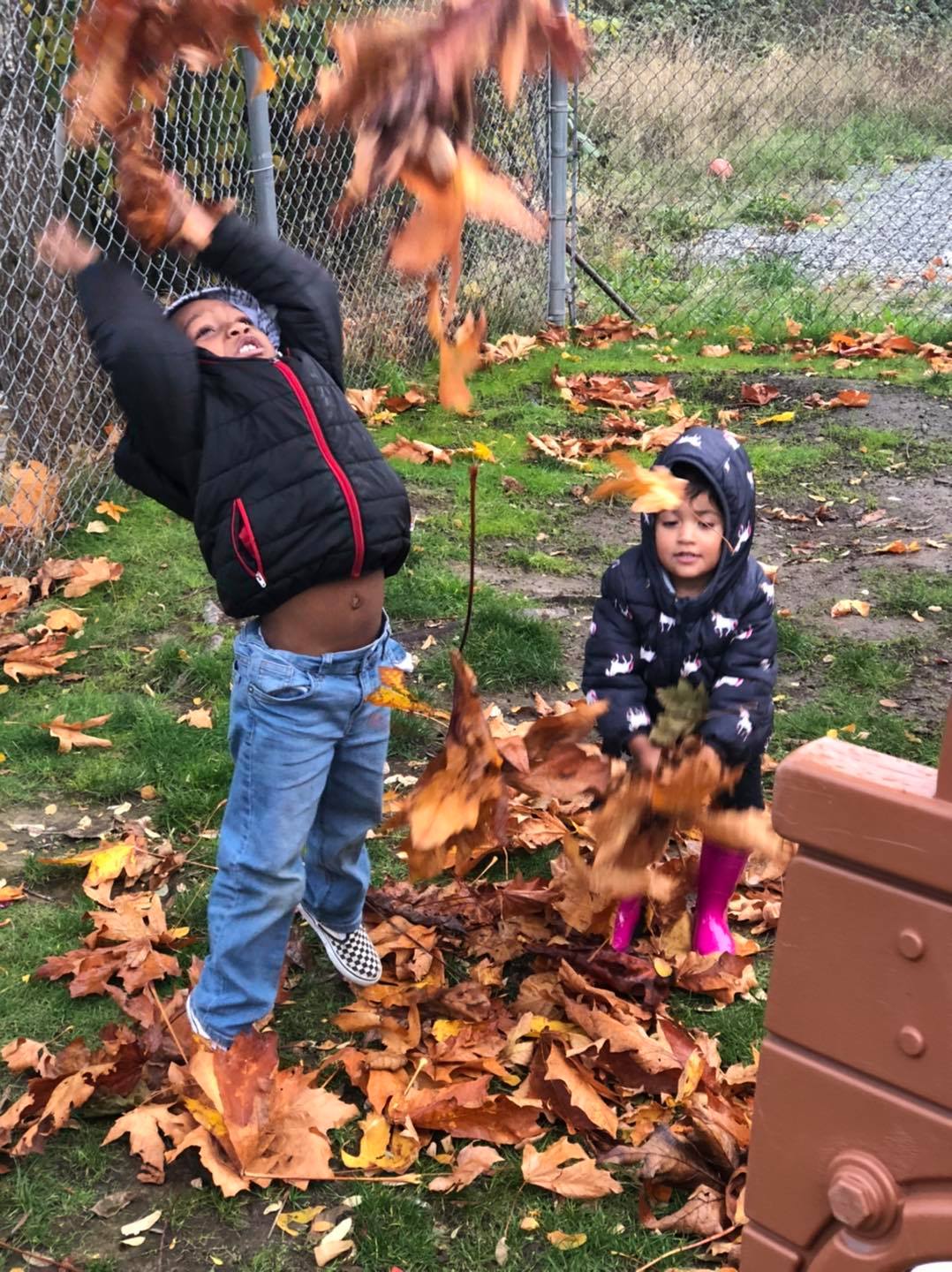 Two children playing in fallen leaves outdoors, one throwing leaves up in the air.