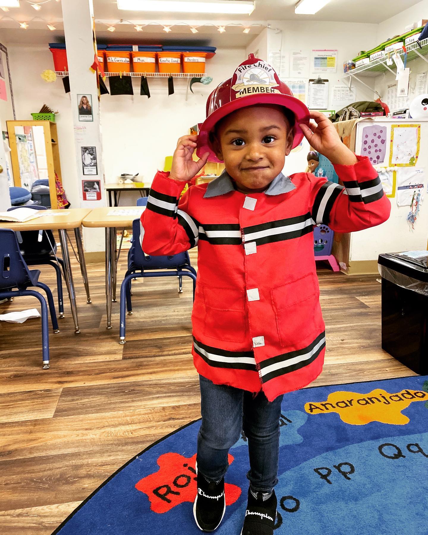 Young child in a firefighter costume, indoors, holding a red helmet.