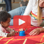 Baby and adult playing with a blue toy on a red mat. The adult wears a rainbow-striped shirt.
