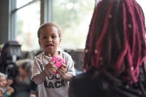 Young child with a pink toy faces forward, speaking to person with pink-streaked dreadlocks. Indoors.