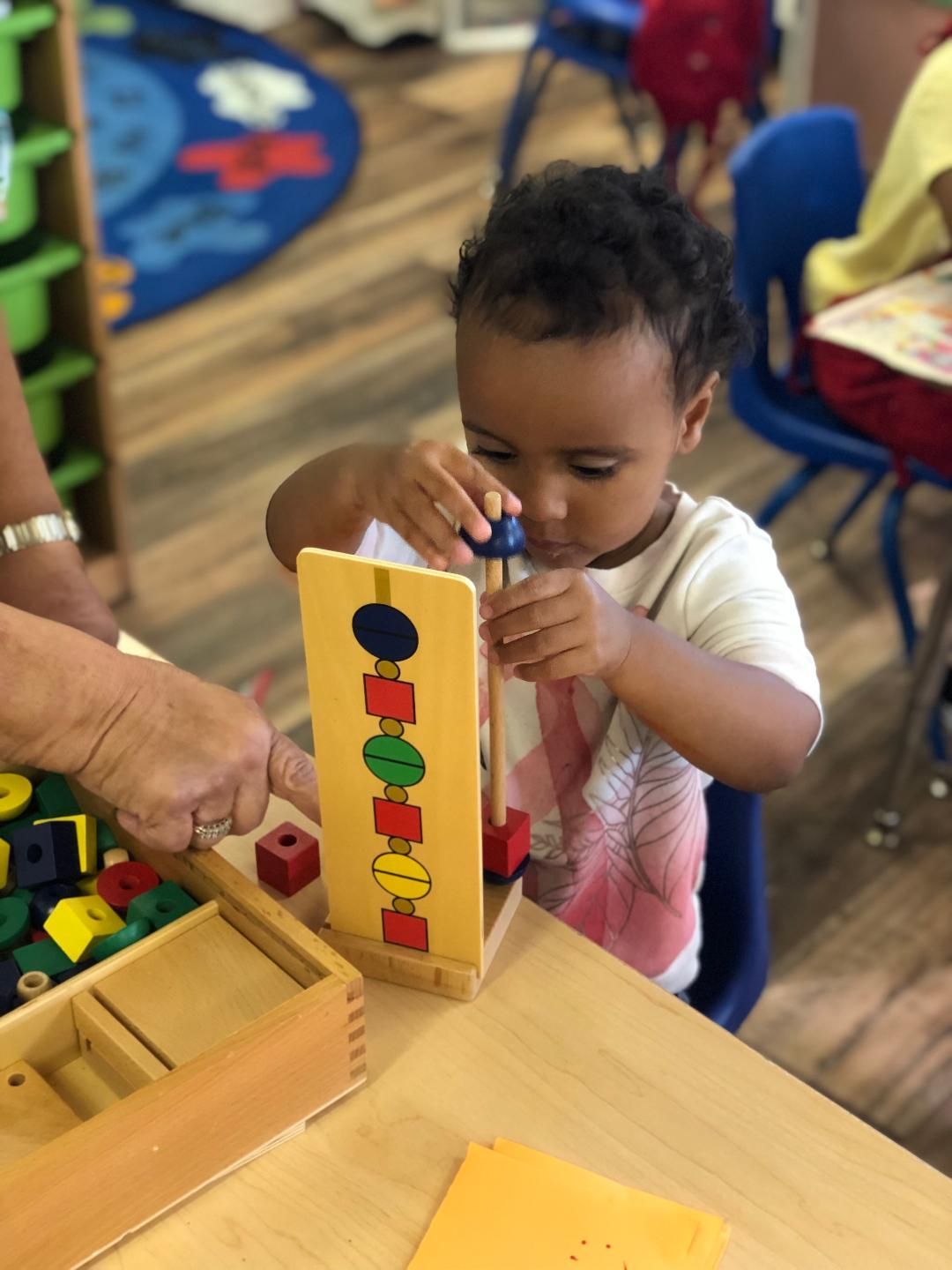 Young child playing with a wooden shape sorting toy at a table in a classroom.