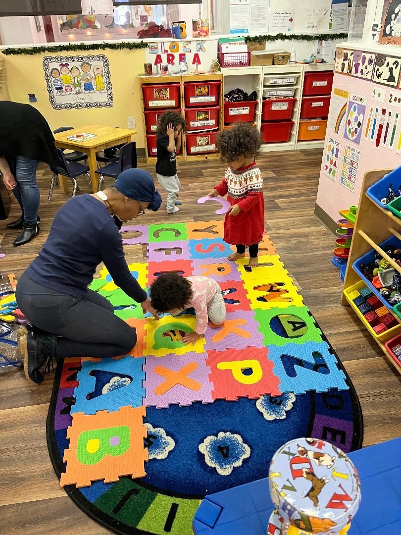 Teacher and toddlers on alphabet floor mat in a brightly colored preschool classroom.
