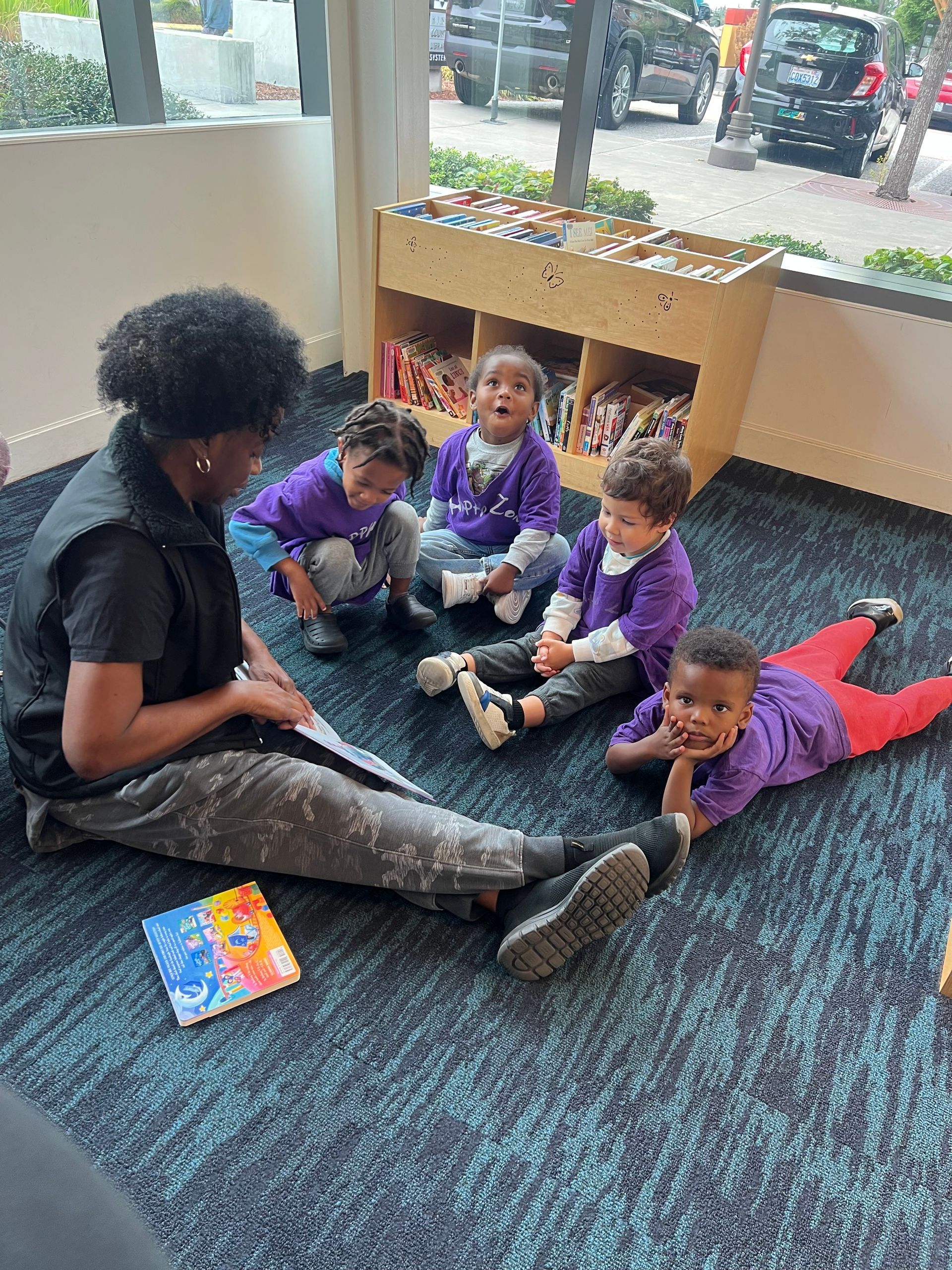 A woman reads to four children in a playroom. They wear purple shirts, smiling.