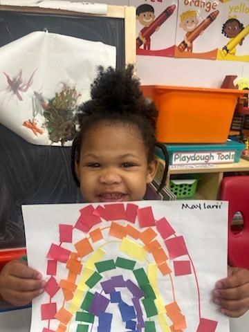 Girl with curly hair holding a colorful paper rainbow in a classroom.
