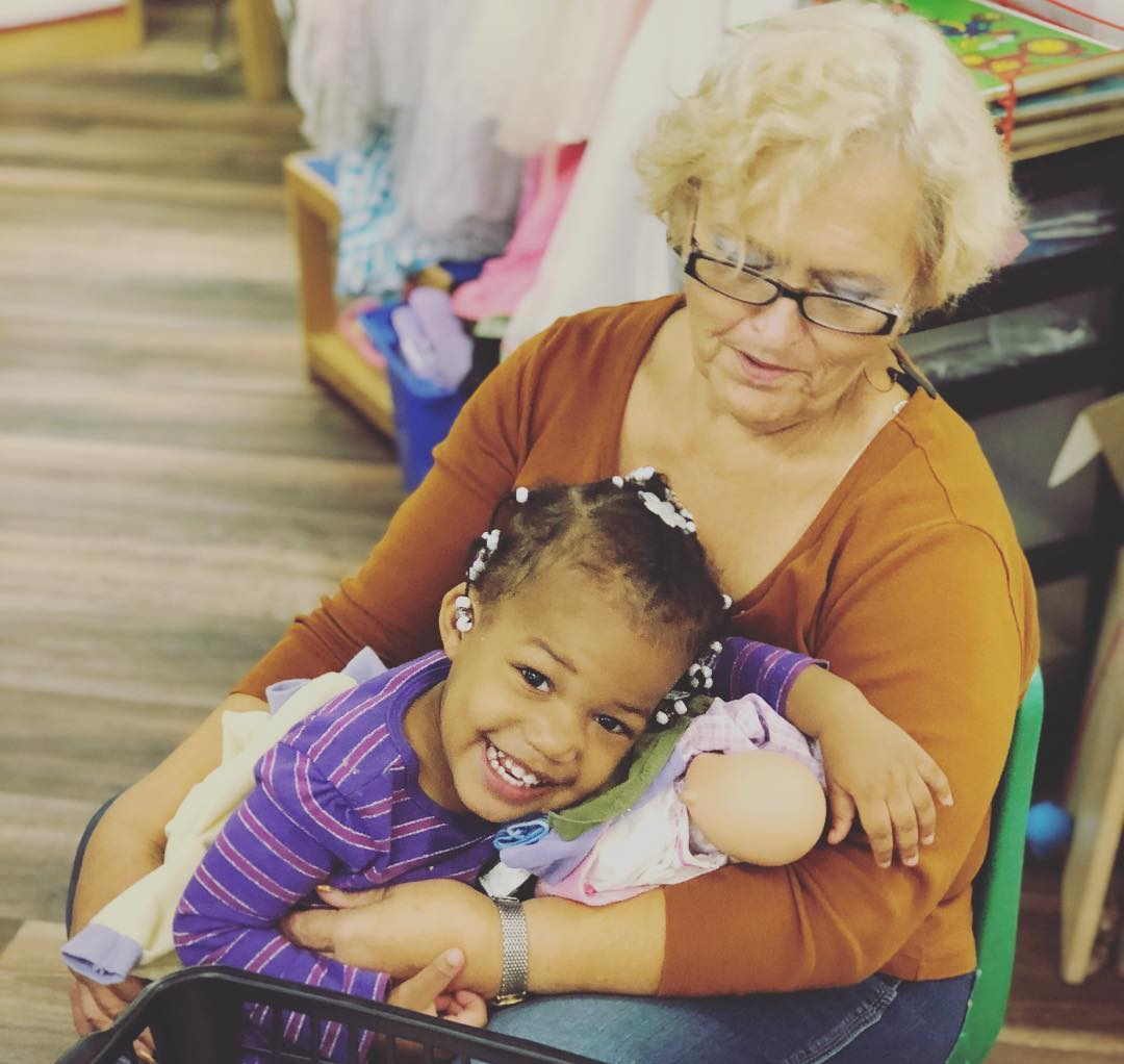 An older woman hugs a smiling Black child holding a doll. Both are indoors.