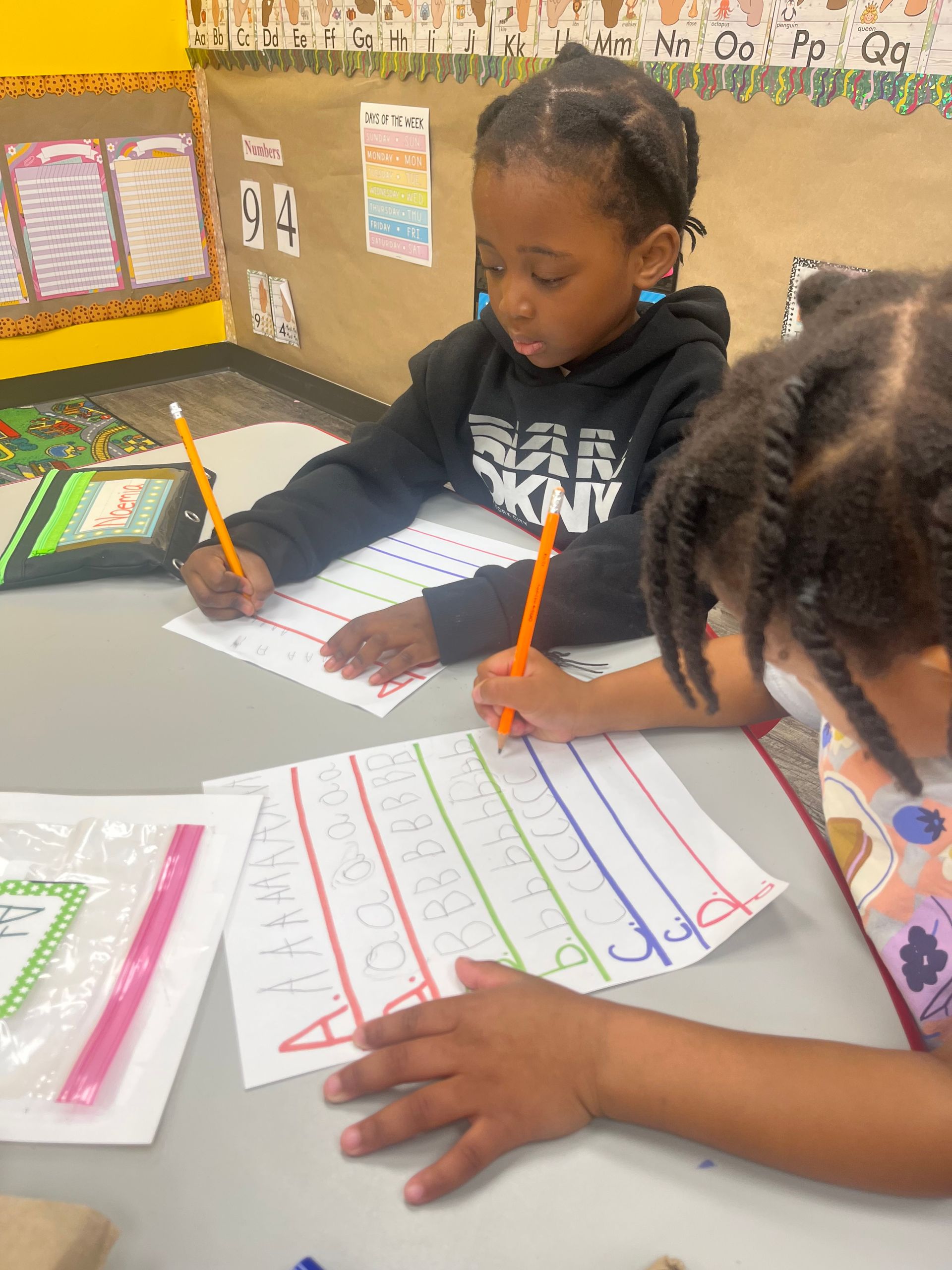 Two young students writing with pencils at a desk, in a classroom setting.