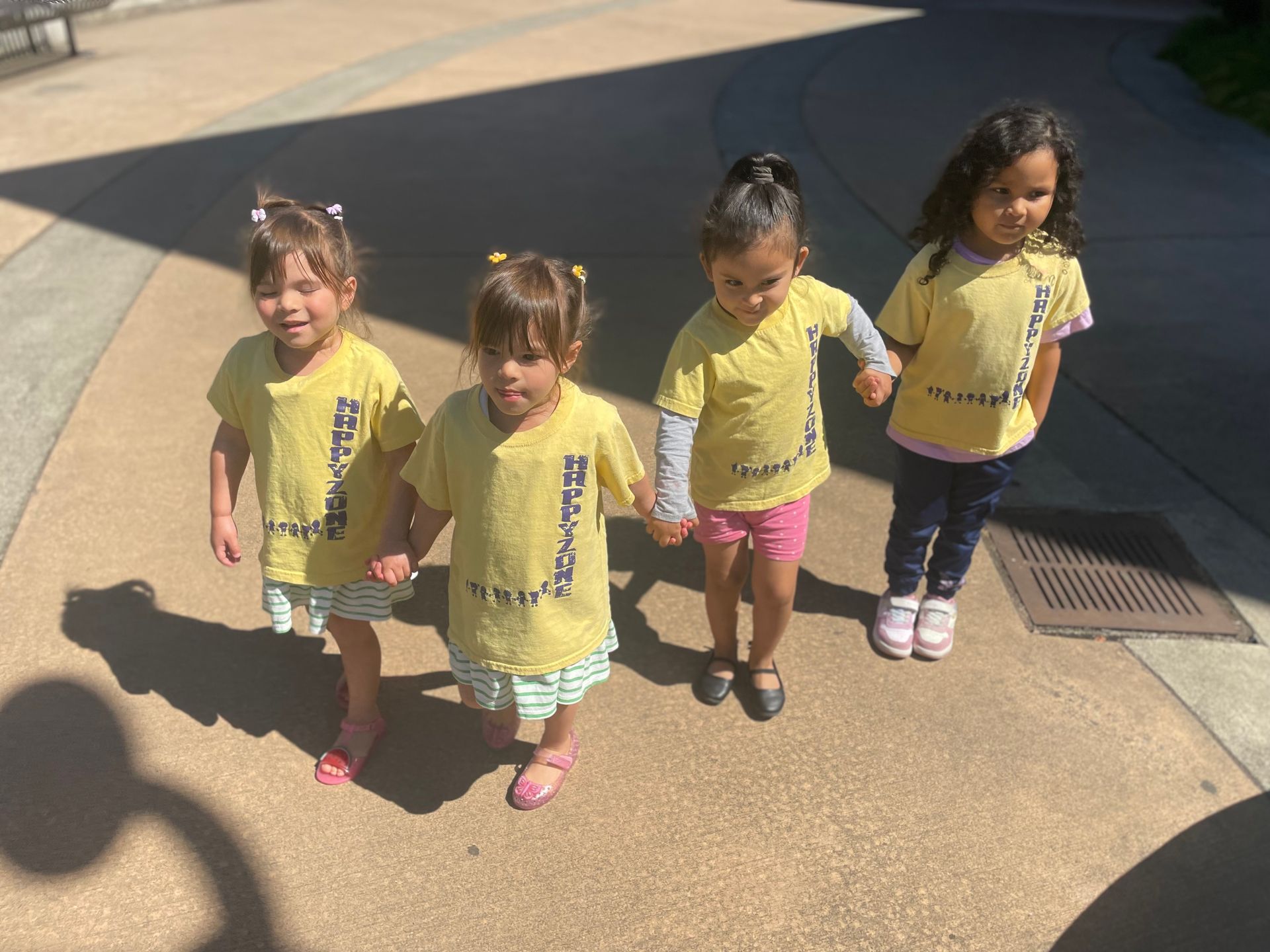 Four young children holding hands, wearing yellow shirts, and standing outdoors.