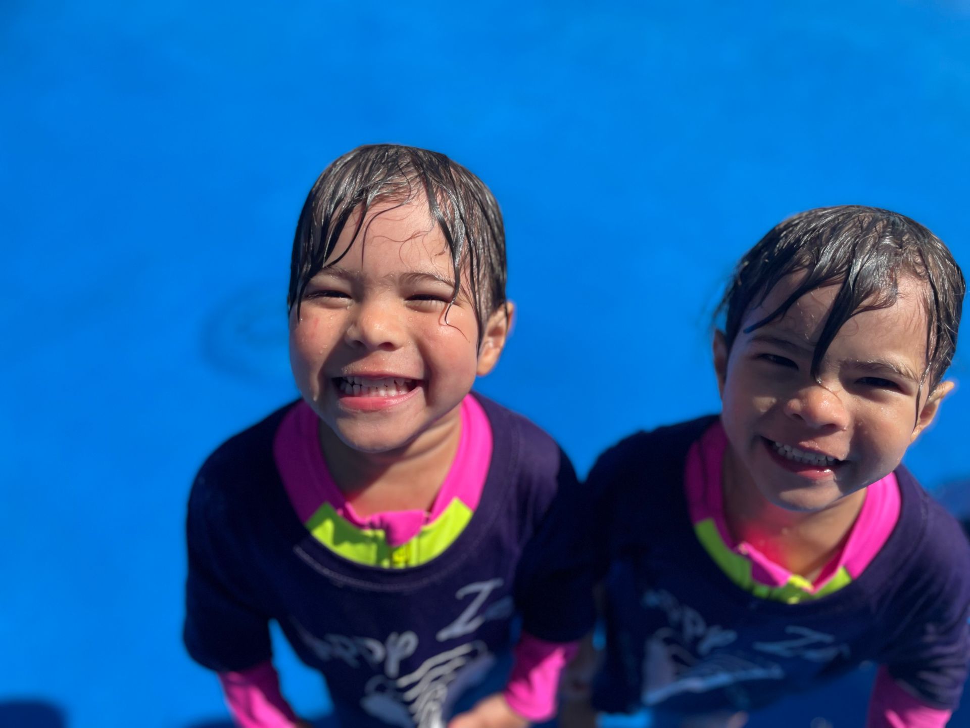 Two young girls smiling, wet hair, wearing swim shirts, in a blue pool.