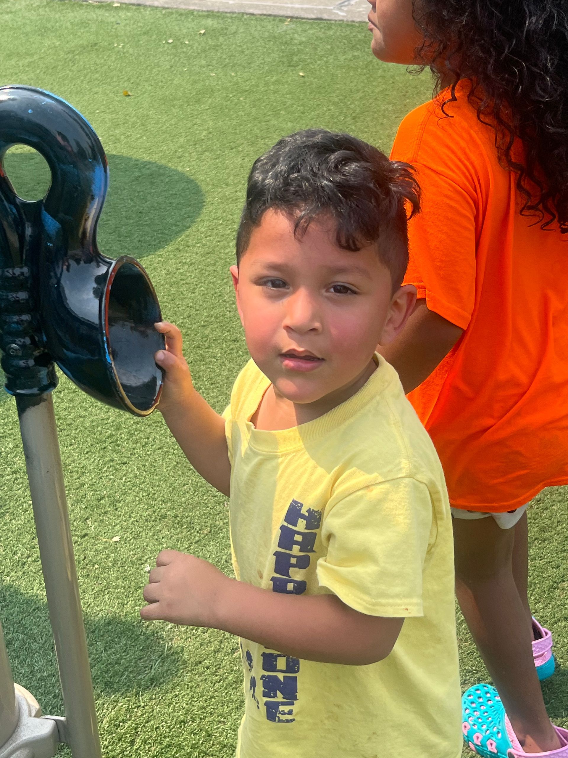 Boy in yellow shirt at playground, holding a black structure. Another person in orange shirt visible.