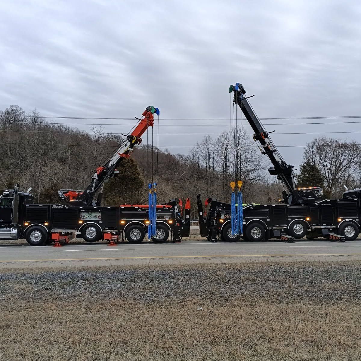Two black tow trucks with booms extended, parked on a roadside with bare trees and a cloudy sky.