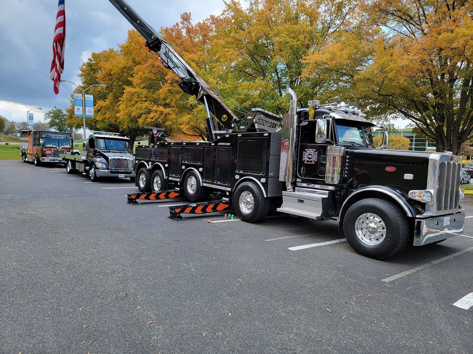 Black tow truck parked on asphalt, other trucks in line; fall foliage in background.