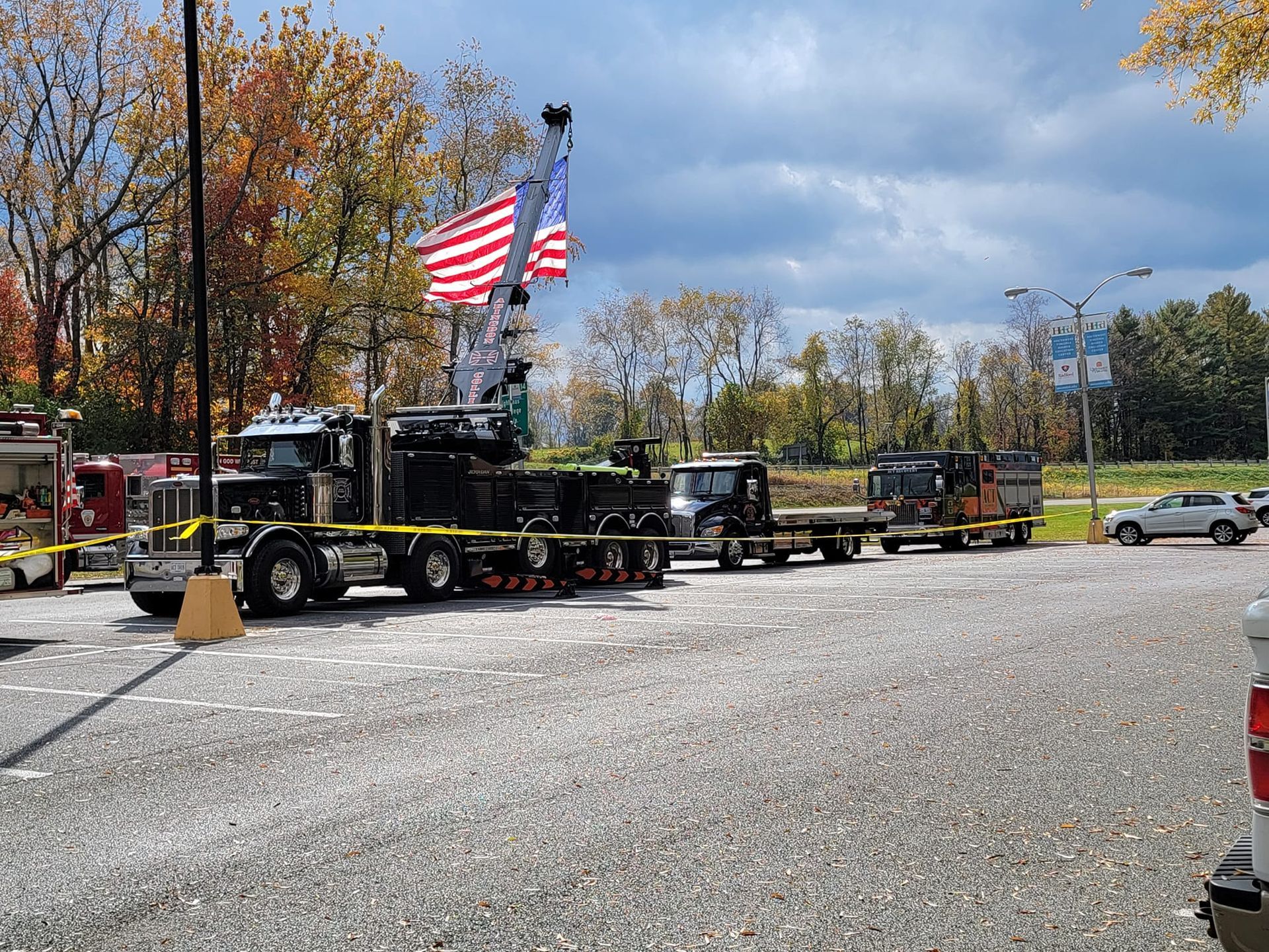 Several large tow trucks and flatbeds parked in a lot, yellow caution tape, American flag in background.