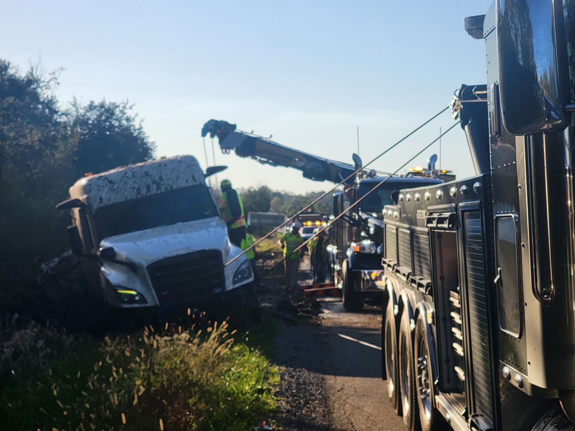 A tow truck pulling a white semi-truck out of a ditch alongside a road.