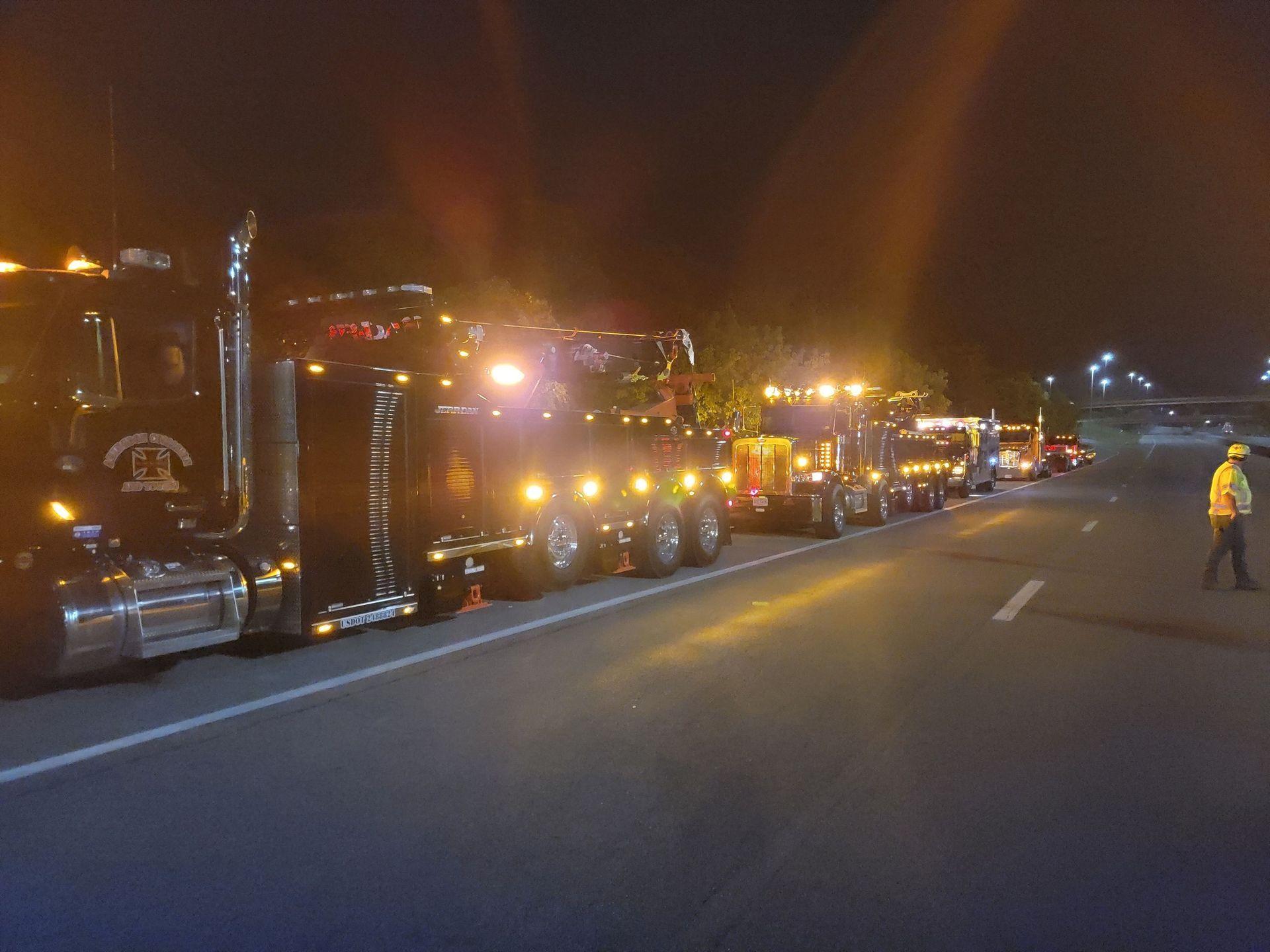 Tow trucks lined up on a road at night with flashing lights, a worker in a safety vest.