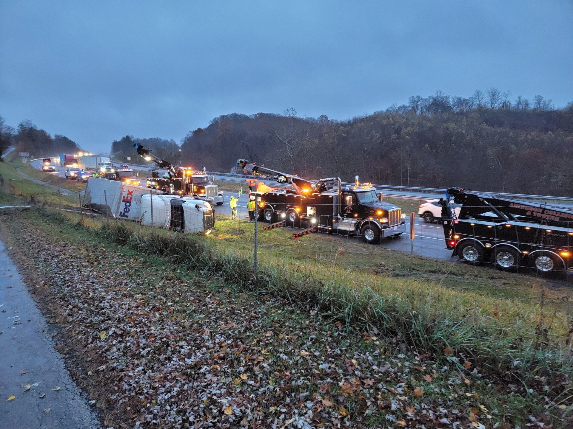 Tow trucks at accident scene on a highway shoulder, with overturned semi-truck and debris, under an overcast sky.
