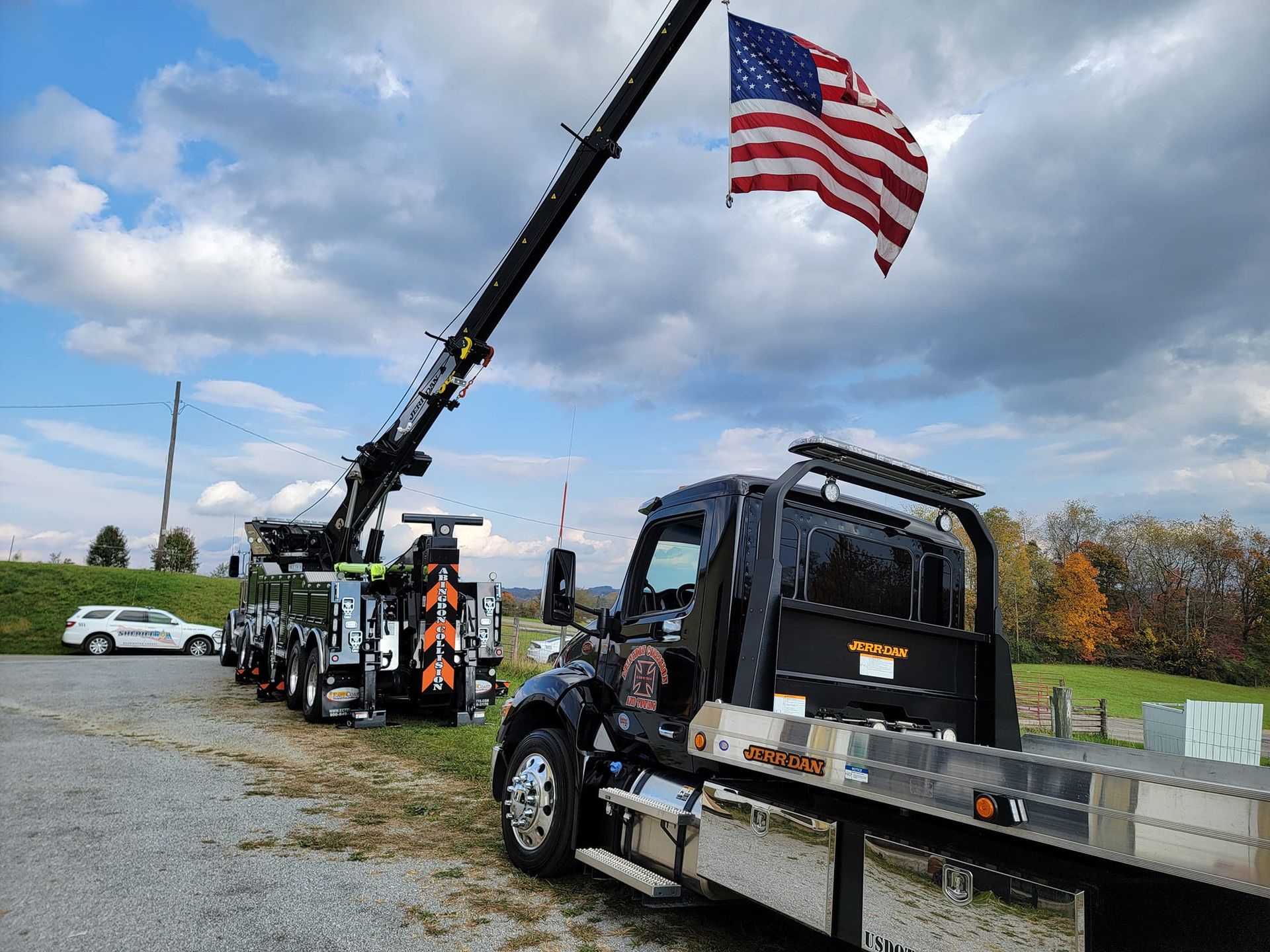 Tow truck with US flag extended, parked in front of field under cloudy sky.