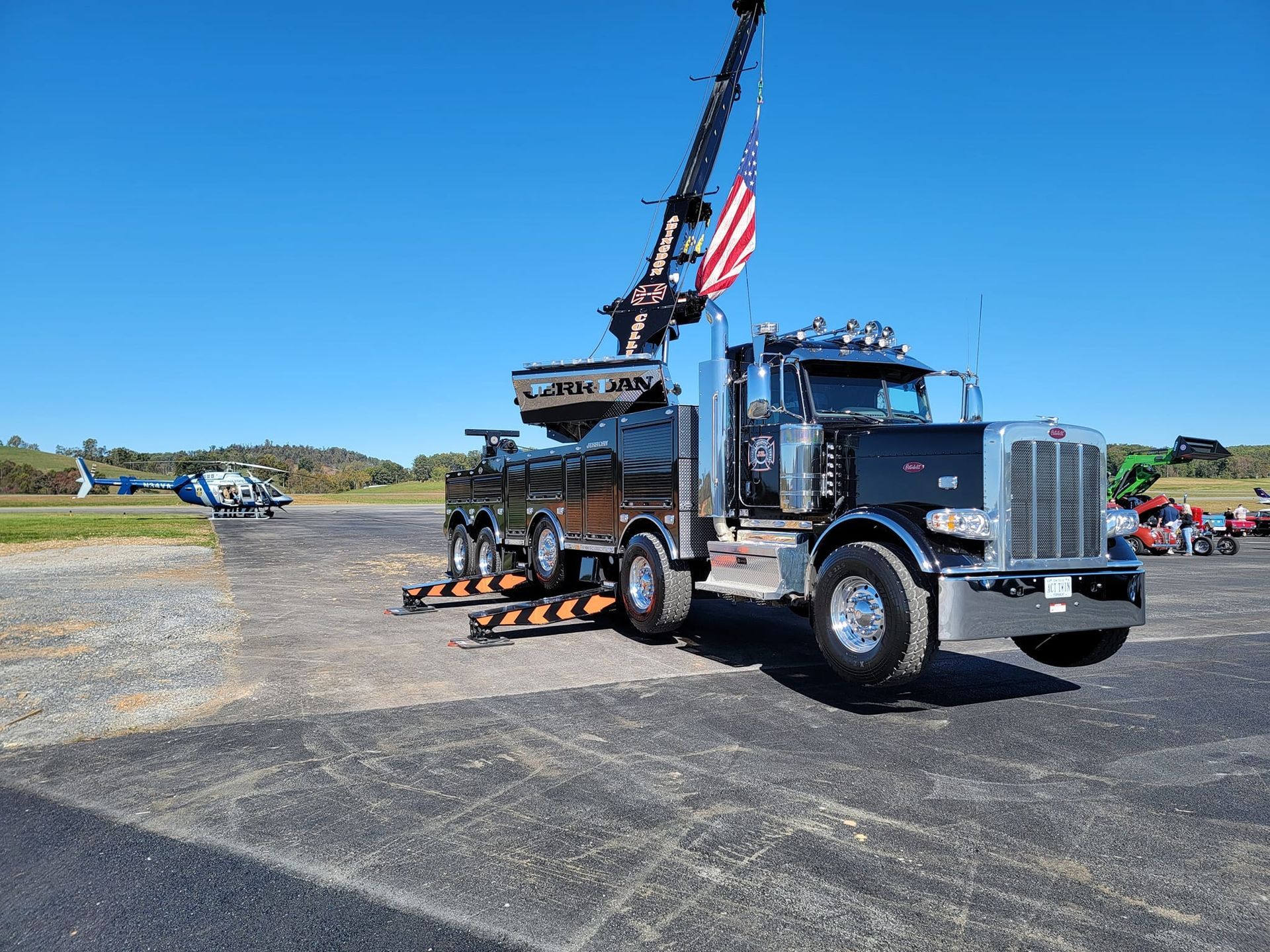 Black tow truck with crane arm and US flag on display, on a paved area with a helicopter and green fields in the background.