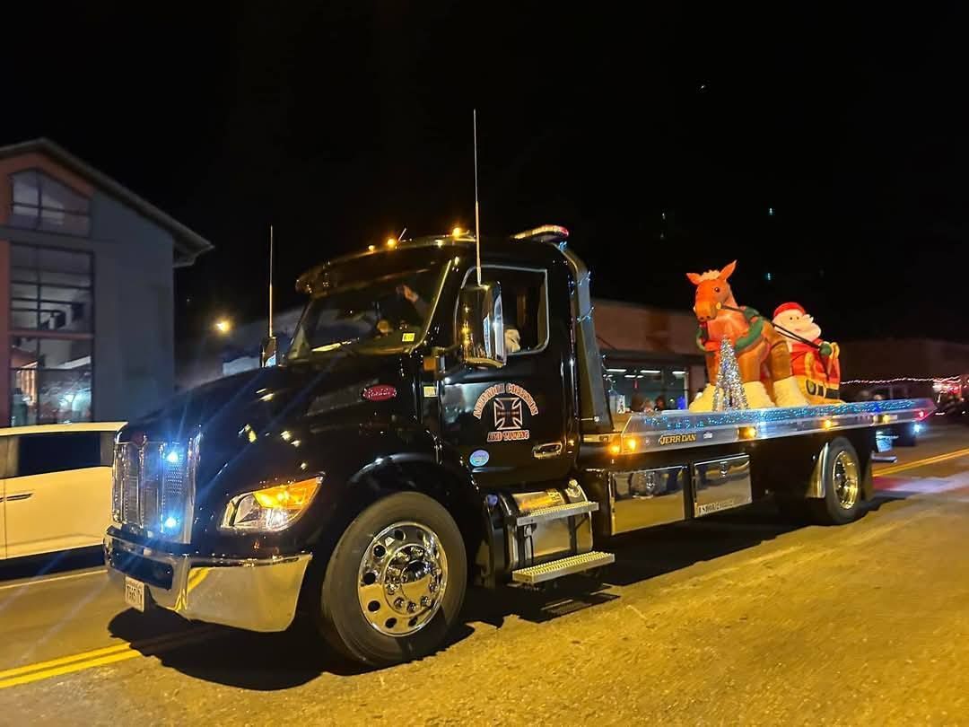 Black tow truck with a decorated reindeer on a flatbed, driving at night.