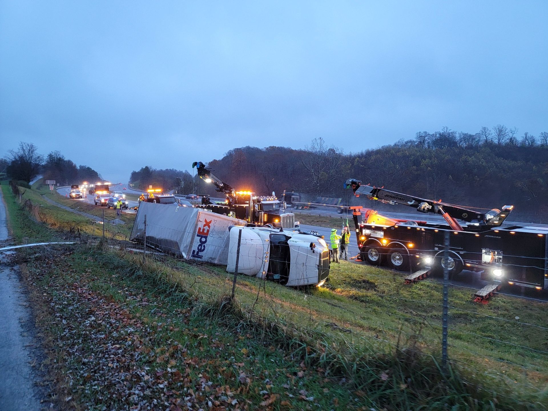 Overturned FedEx truck on a roadside, tow trucks, flashing lights, recovery operation underway on a cloudy day.