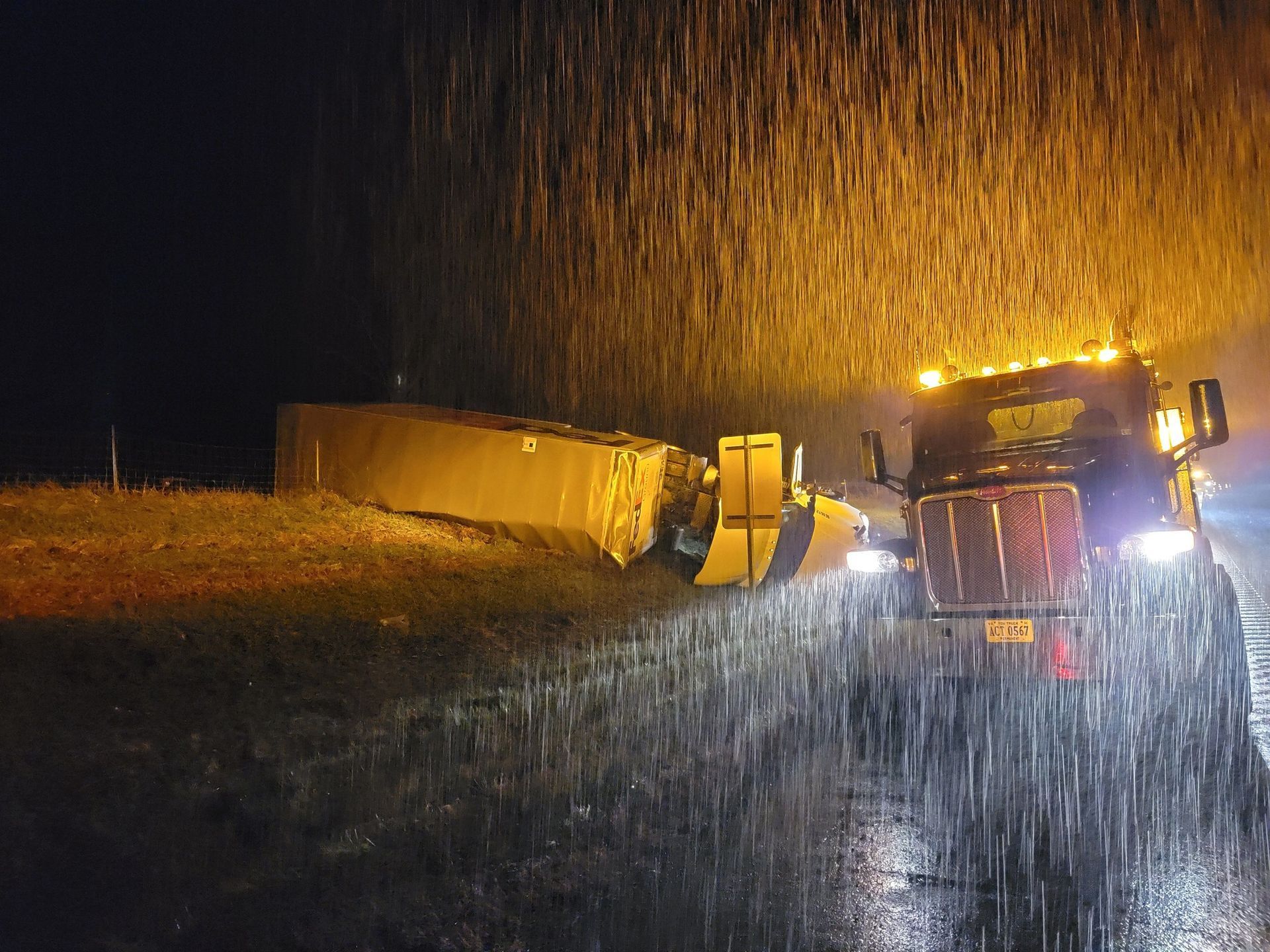 Semi-truck off the road at night in rain, headlights on, trailer damaged.