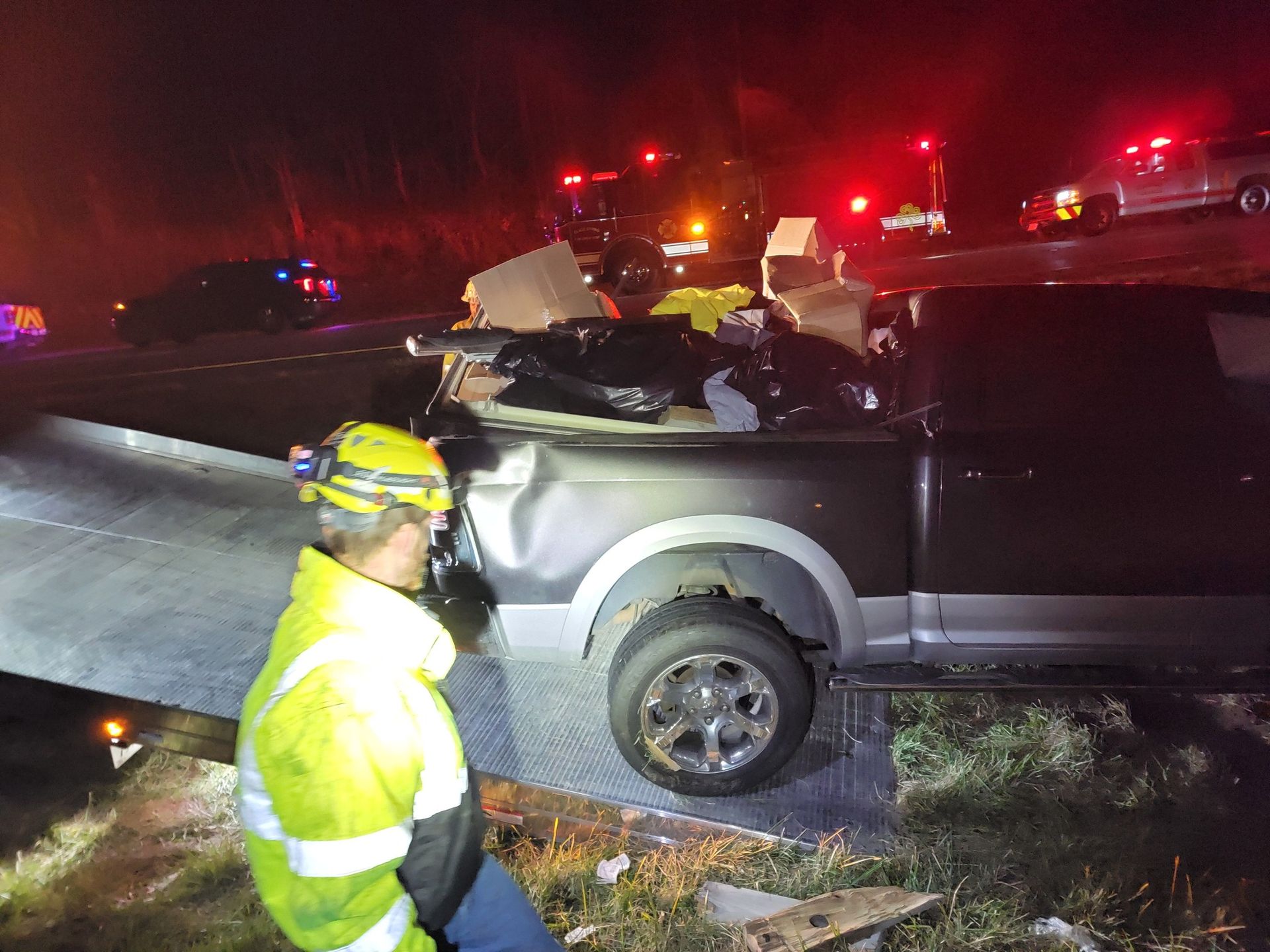 A damaged pickup truck at a nighttime accident scene; emergency vehicles with flashing lights are present. A person in a yellow vest examines the truck.