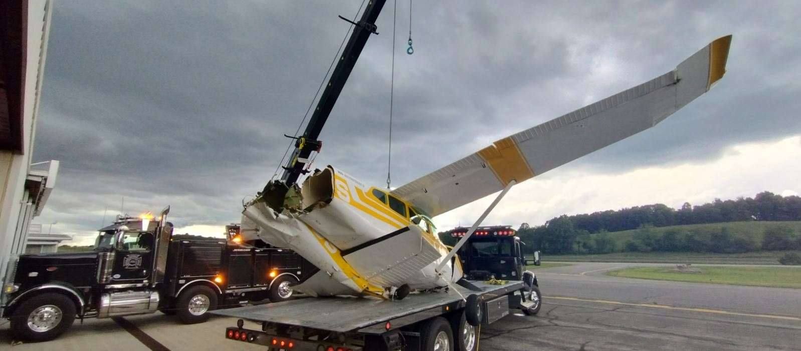 A small airplane being lifted by a crane onto a flatbed tow truck at an airport under a cloudy sky.