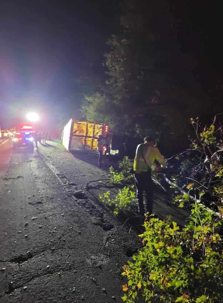 A truck overturned on a dark road next to a treeline; emergency responders are on the scene with flashing lights.