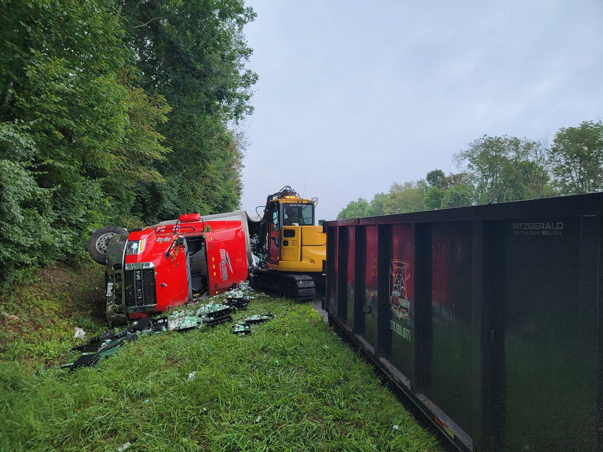 Red overturned truck next to a yellow excavator and a dark boxcar on grass next to trees.