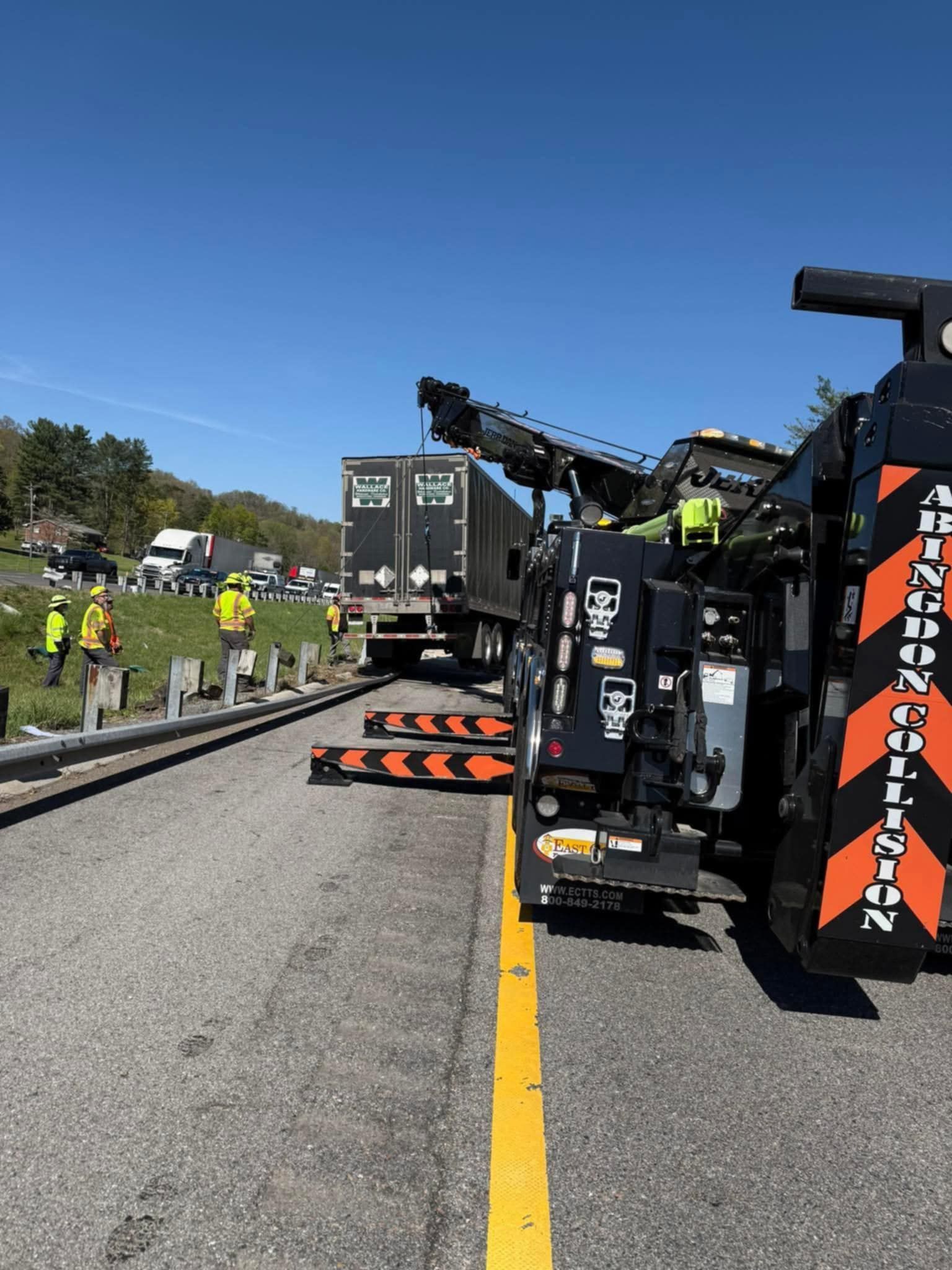 Tow truck pulling a semi-truck from the side of a road, traffic and workers visible in the background.