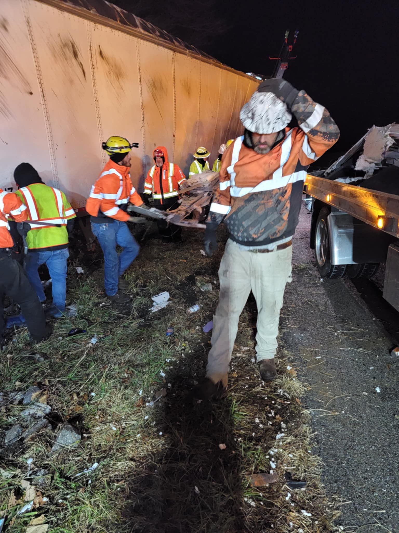 Emergency responders at night near a damaged truck trailer, one holding a tool aloft, others helping.