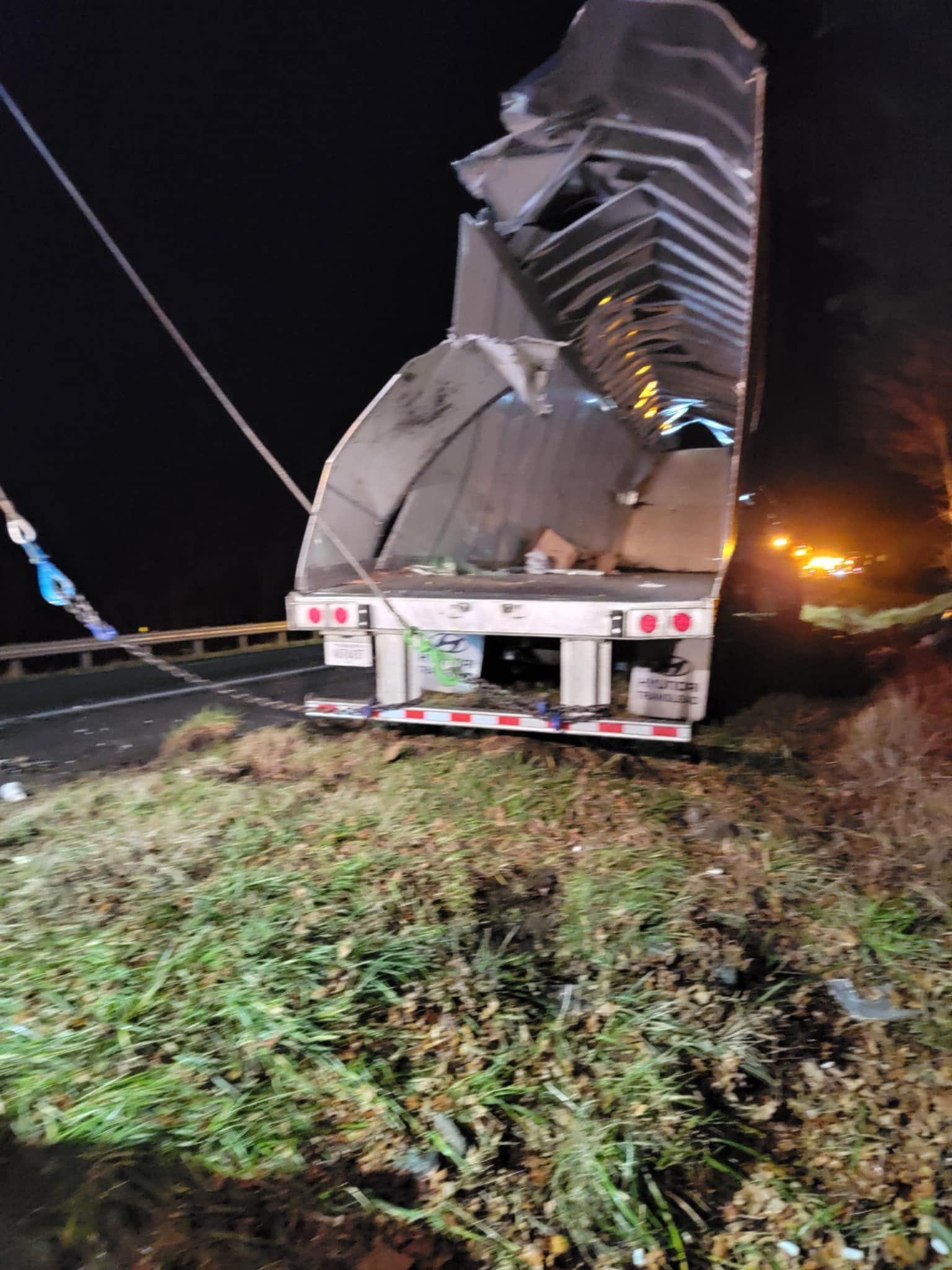 Damaged semi-trailer overturned on roadside at night. Metal roof and sides are heavily damaged.