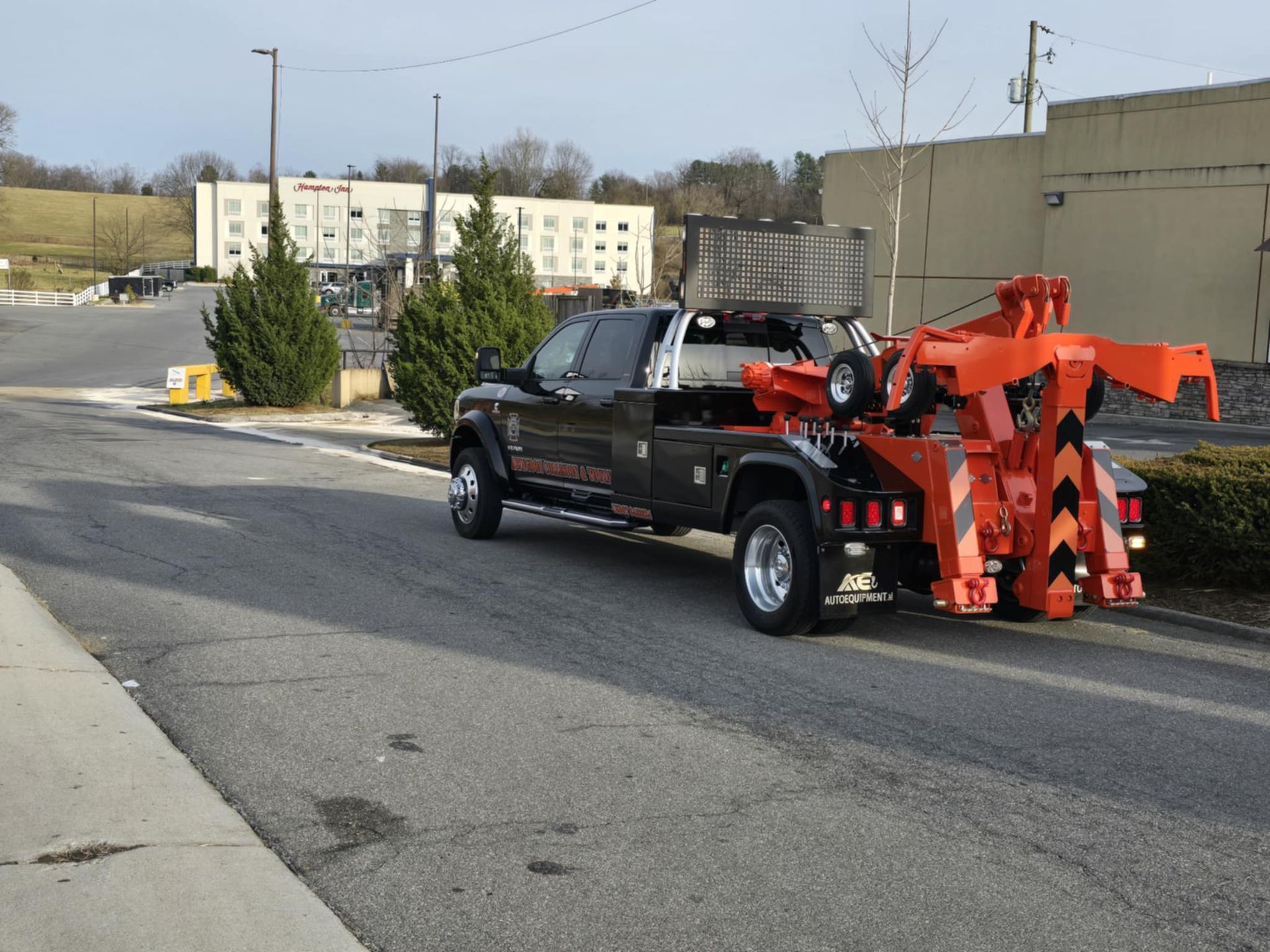 Black tow truck with orange towing equipment on a road near a building and greenery.