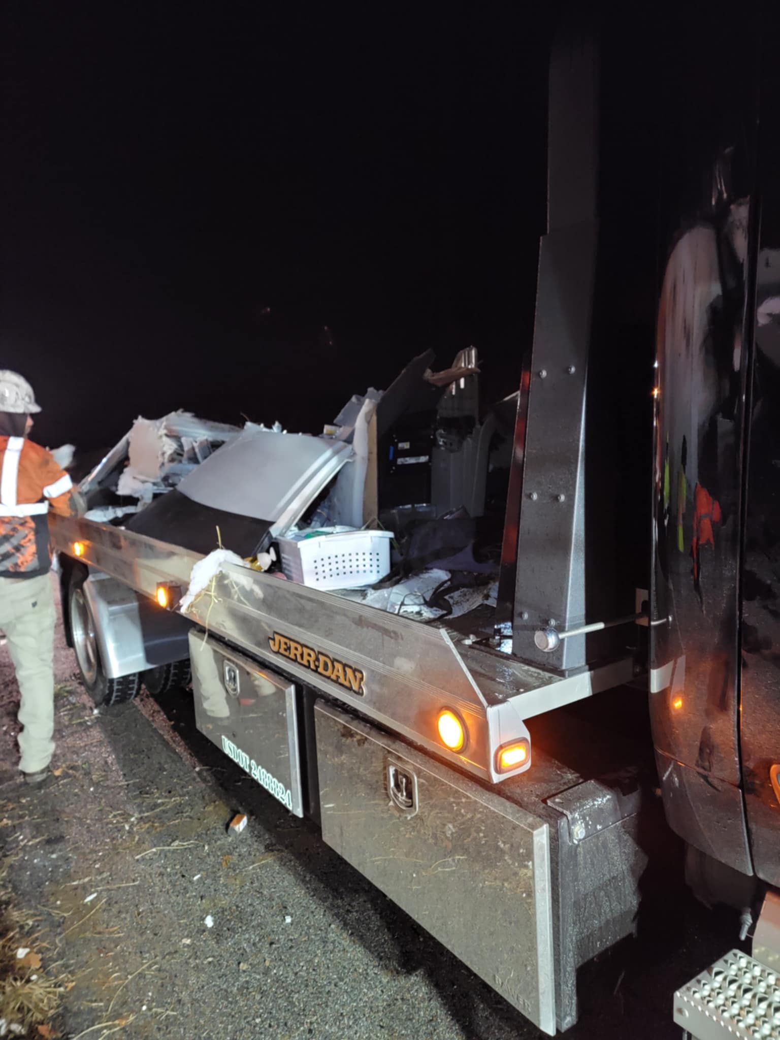 A flatbed trailer loaded with debris next to a semi-truck at night; a person in an orange vest stands nearby.