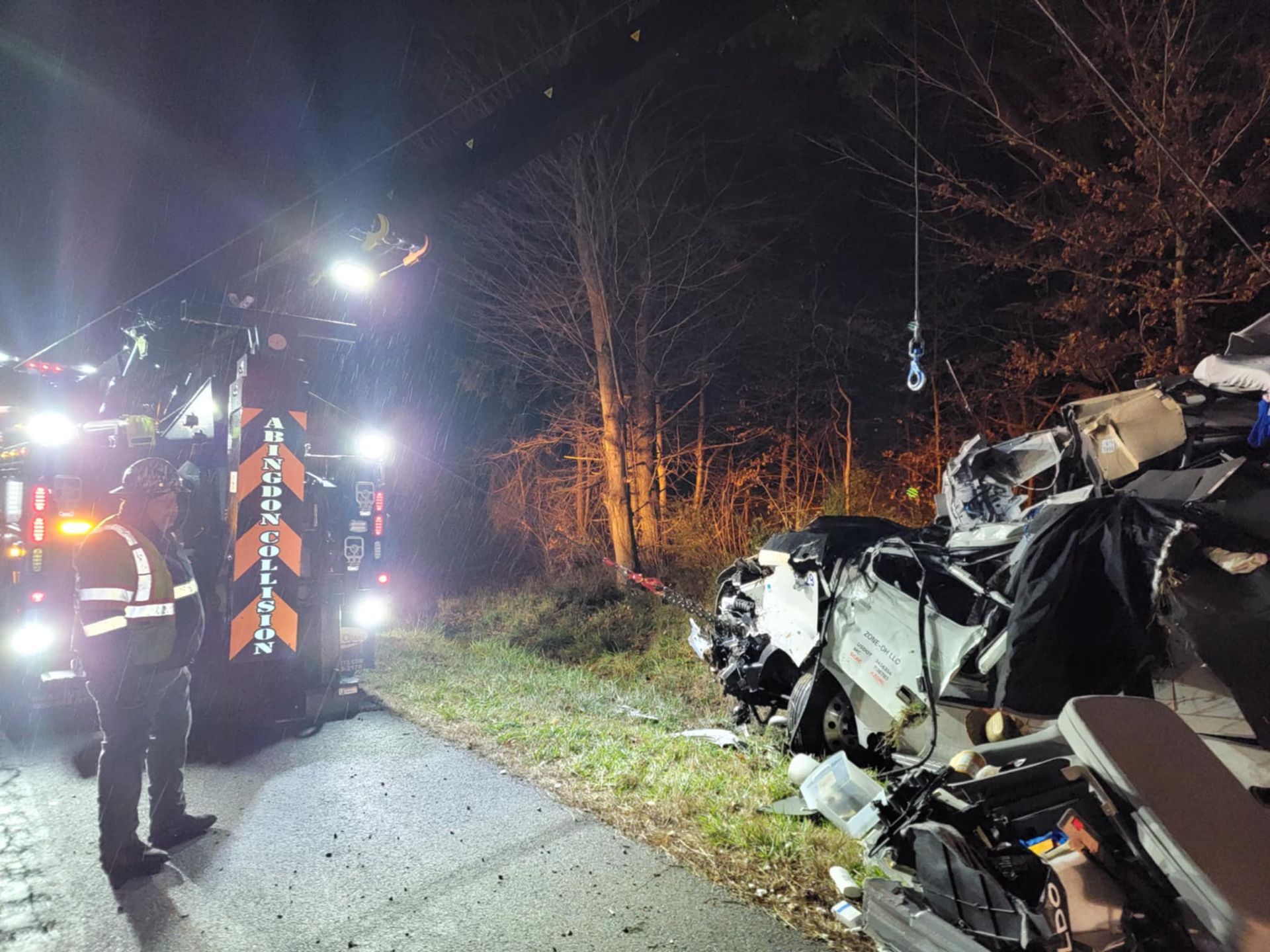 Tow truck at night next to a severely damaged vehicle off the side of a road. A person in a safety vest stands nearby.