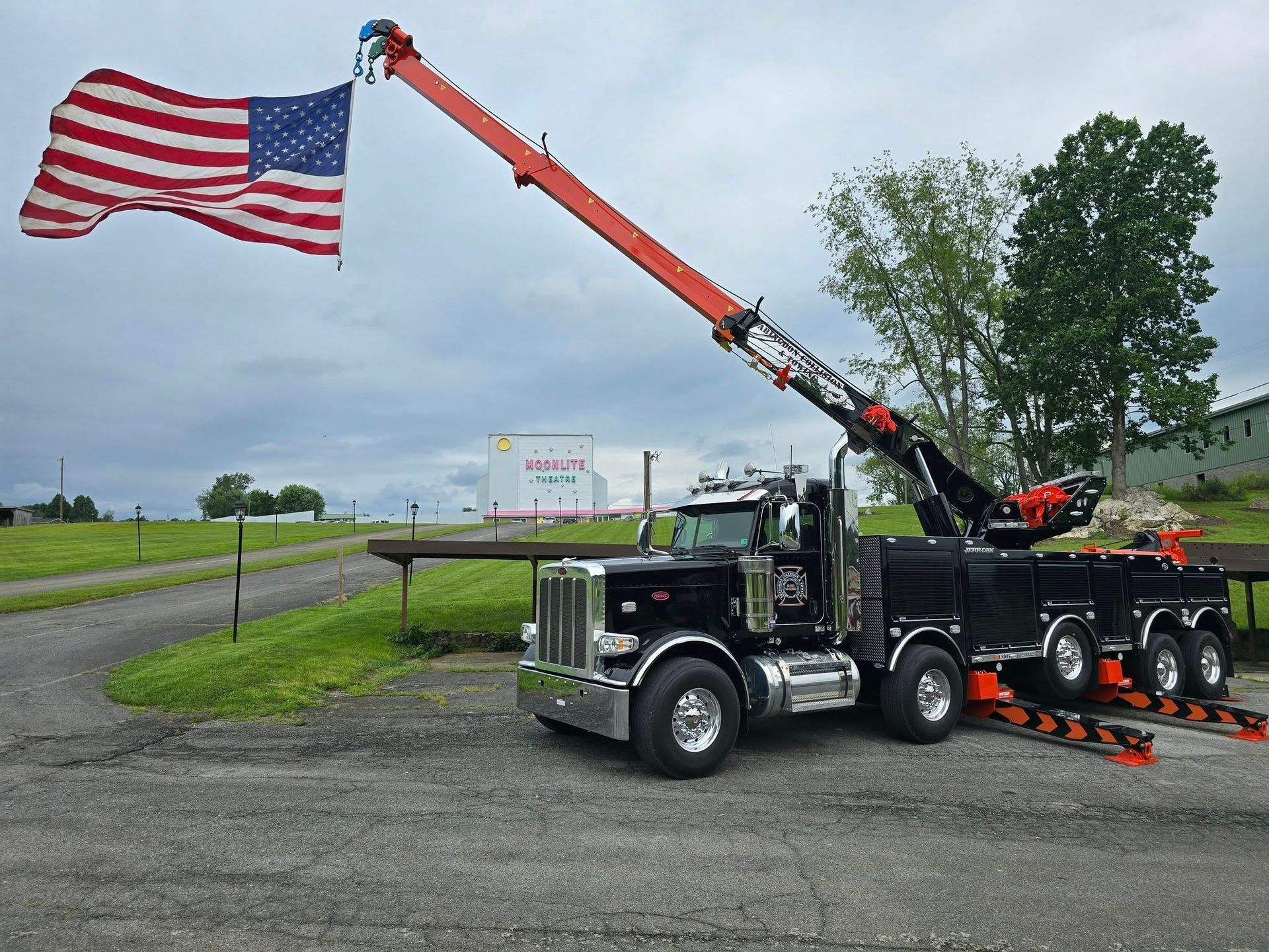 Black tow truck with US flag extended, parked on asphalt. Overcast sky.