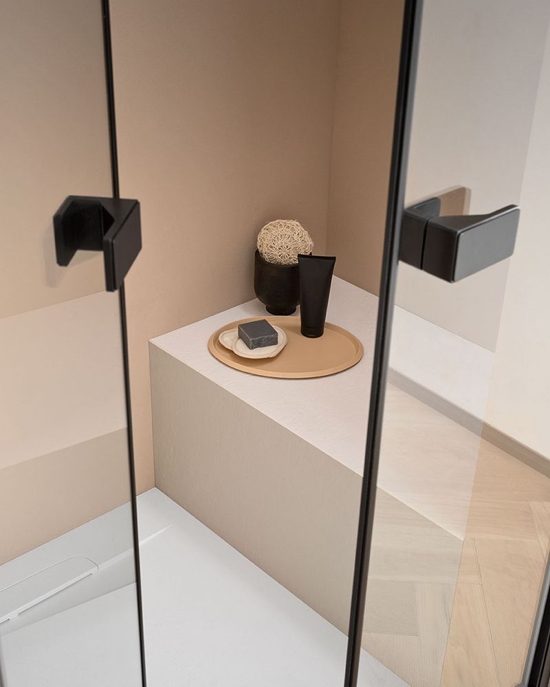 Mensola da bagno minimalista con vassoio, tazza e oggetti decorativi accanto a uno specchio e al box doccia.