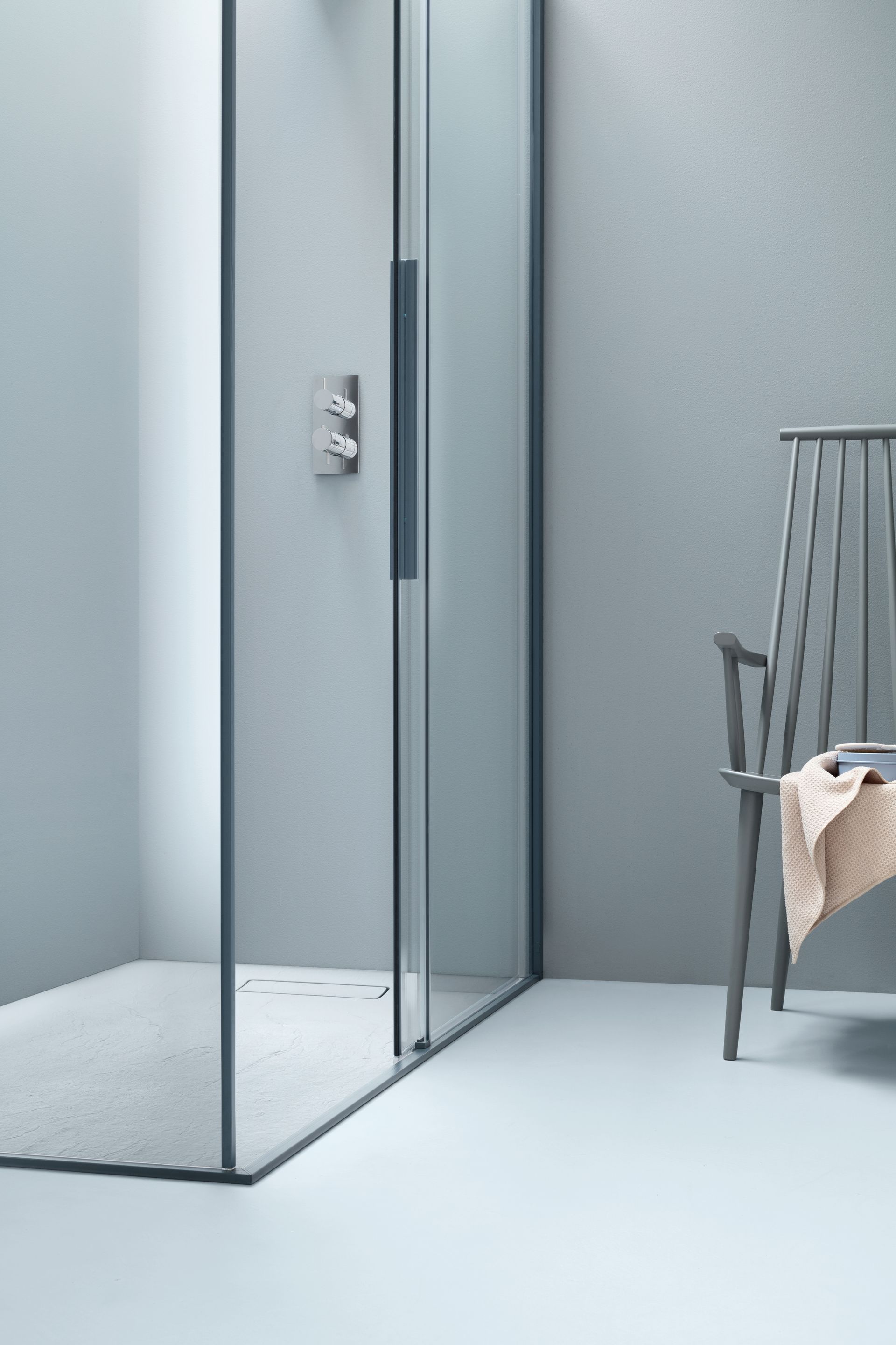Bagno minimalista con porte doccia in vetro, pareti grigie e una sedia con un asciugamano.