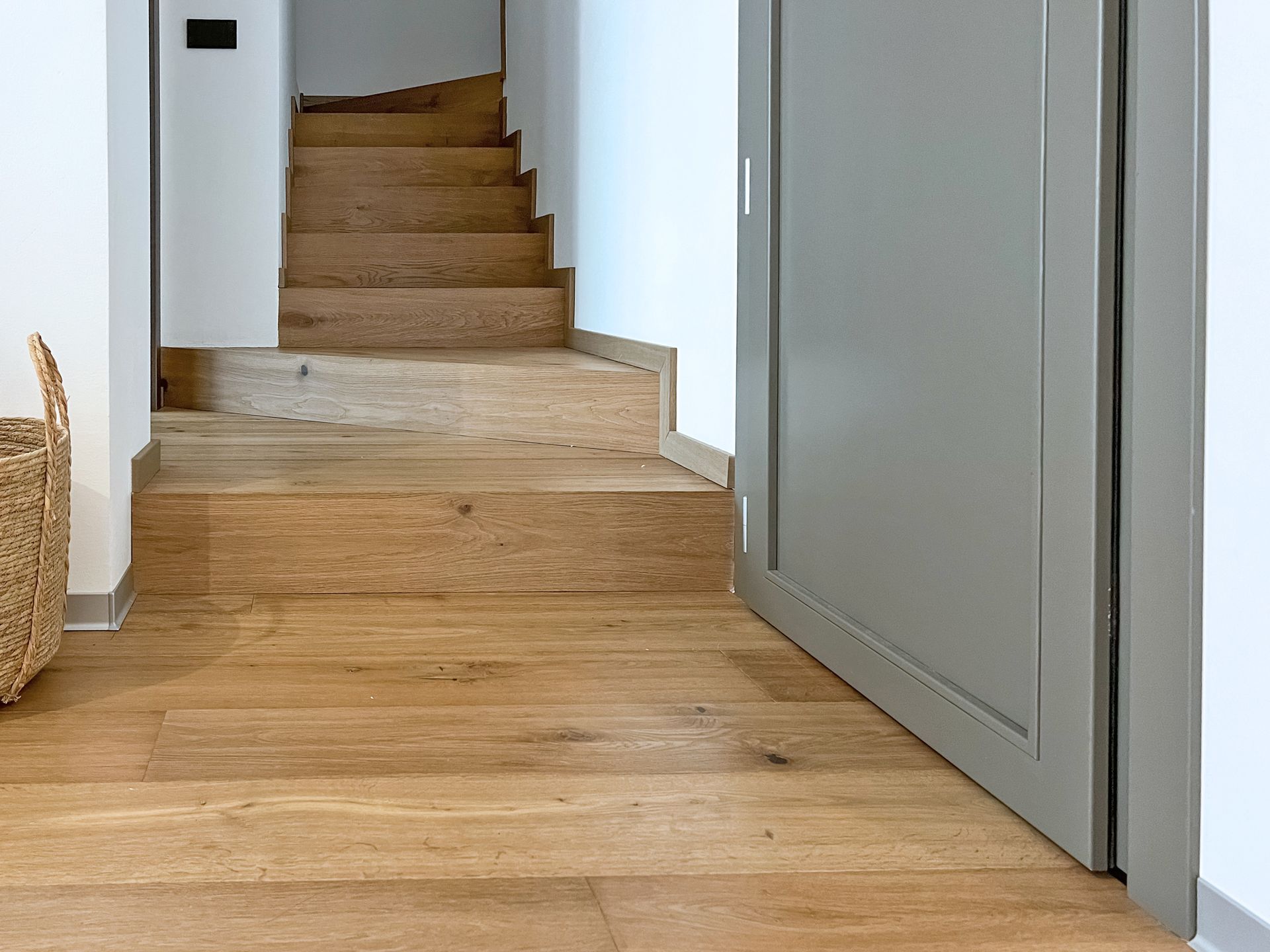 Corridoio con pavimento in legno che conduce a una stretta scala, accanto a una porta grigia parzialmente aperta.